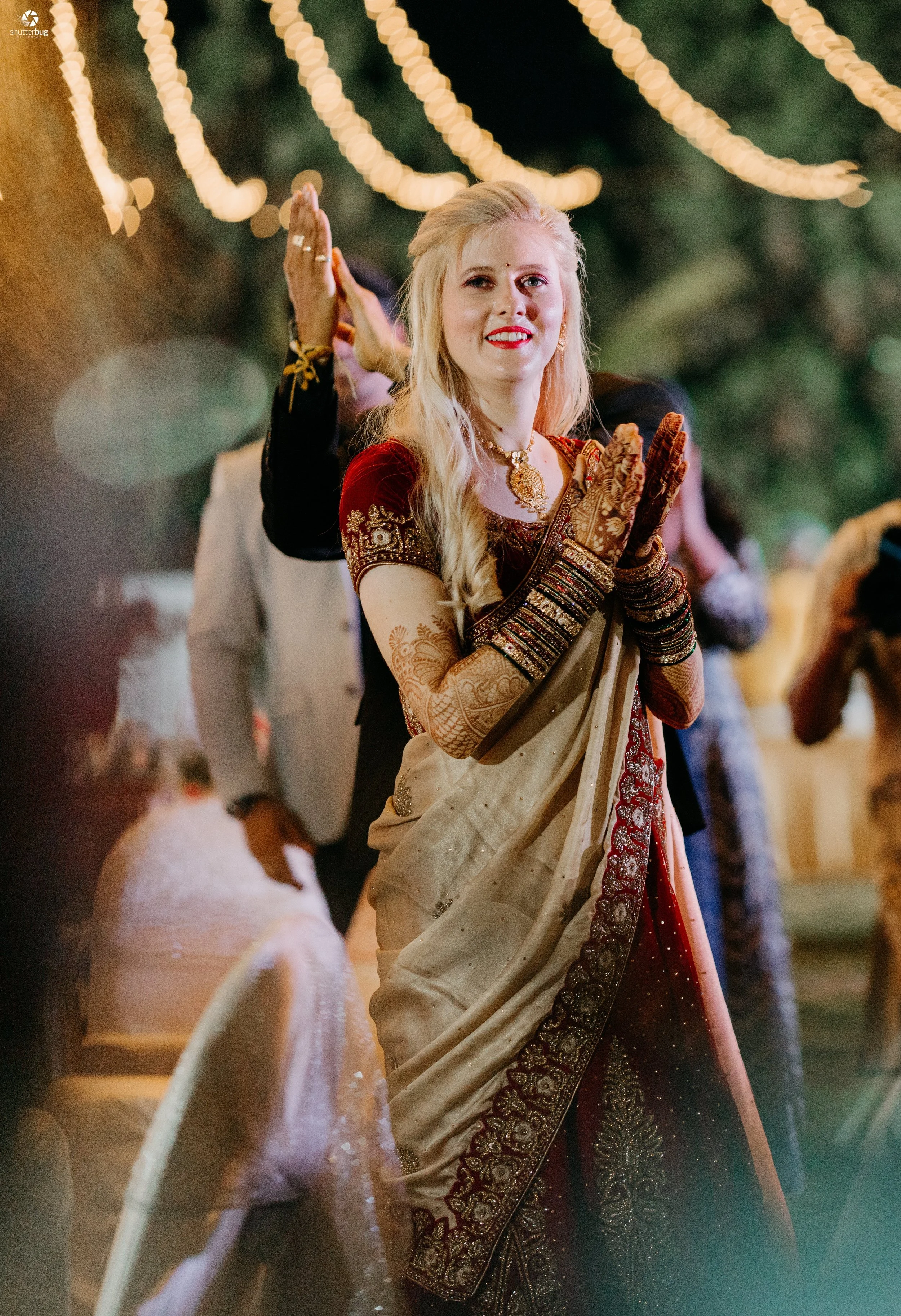 A woman in traditional Indian attire, including a beige and red sari with gold embroidery, is smiling with her hands pressed together in a prayer or greeting gesture at an outdoor event decorated with string lights.