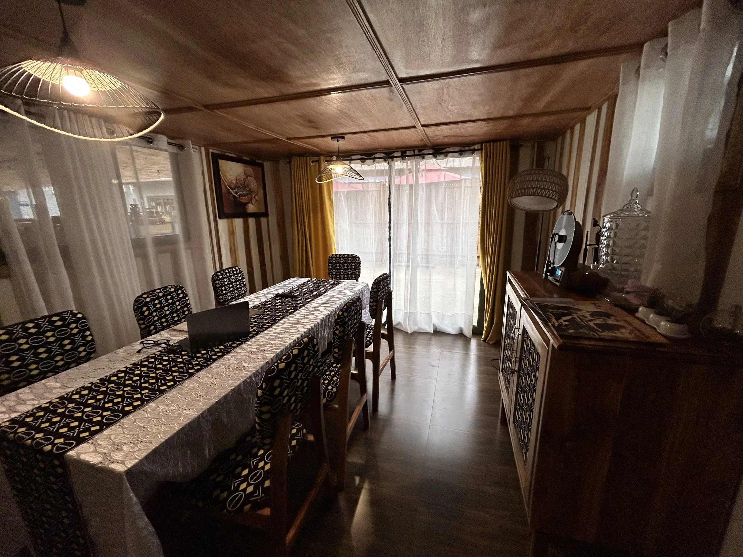 Salle à manger avec une grande table recouverte d'une nappe et des chaises à motif noir et blanc, éclairée par deux plafonniers, avec des rideaux jaunes et blancs aux fenêtres et un buffet en bois avec des décorations.