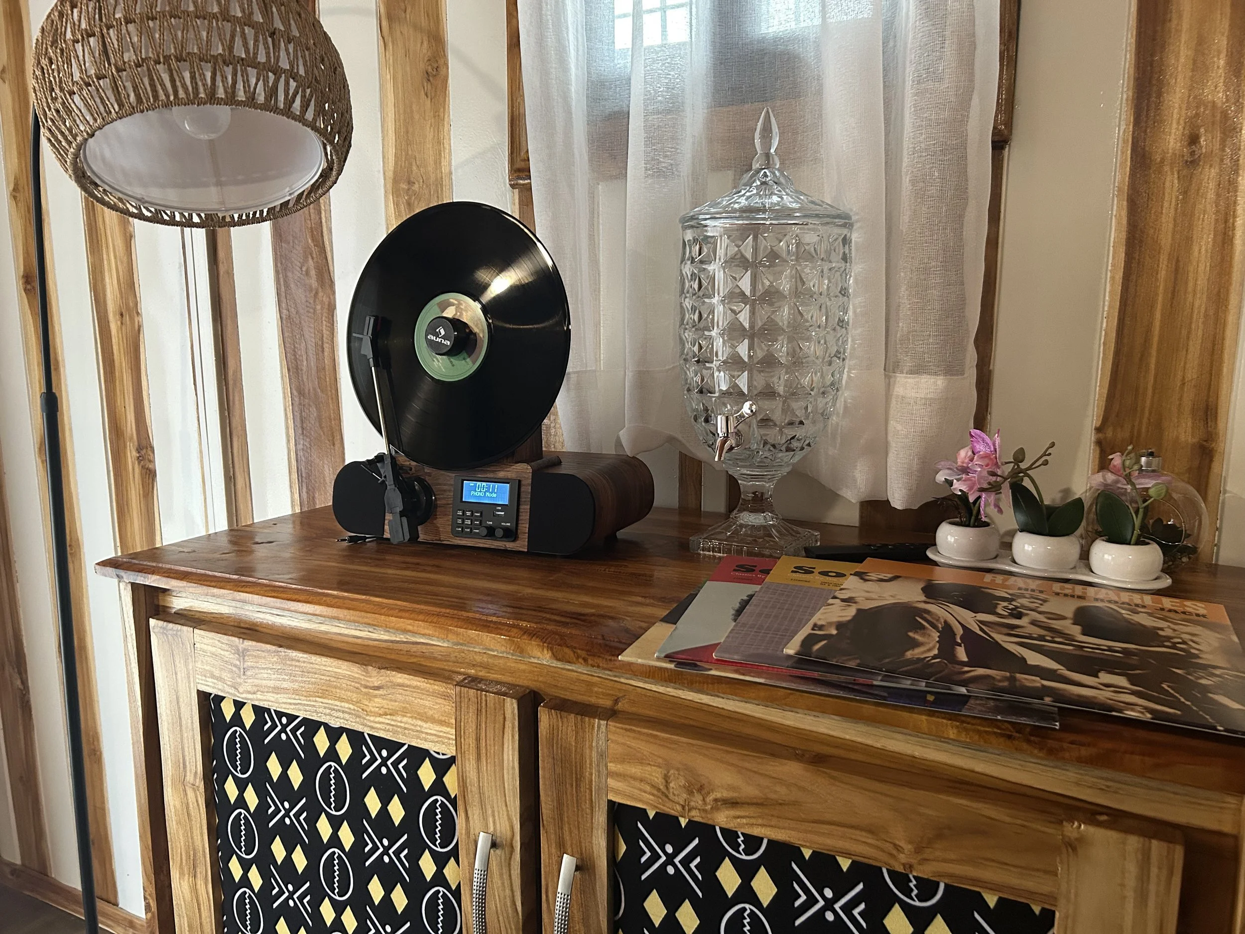 Un meuble en bois avec un tourne-disque, un grand vase en cristal avec robinet, de petites pots de fleurs avec des orchidées, et des magazines.