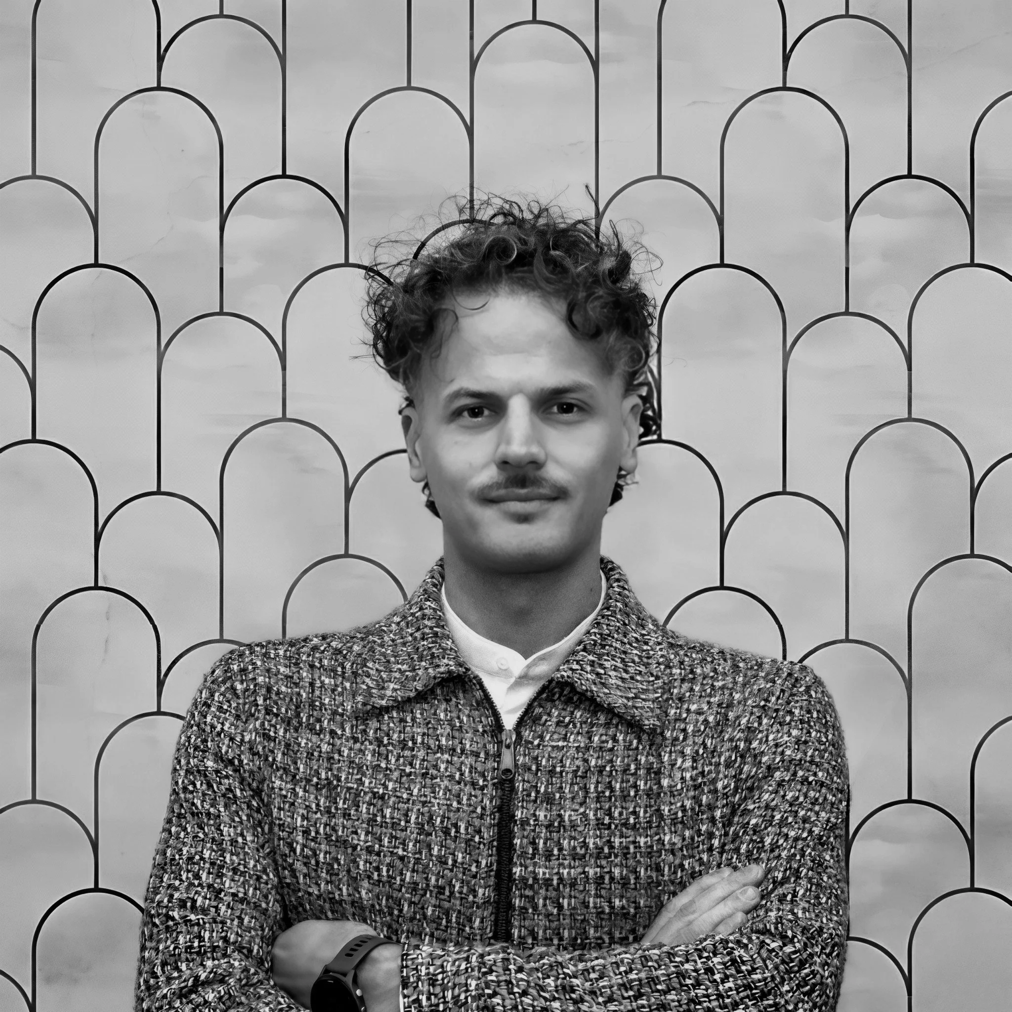 Ritratto di un uomo con i capelli ricci e un pizzetto, vestito con una giacca a TWEED e una camicia bianca, che incrocia le braccia, posizionato davanti a uno sfondo decorativo con figure a forma di archi stilizzati.