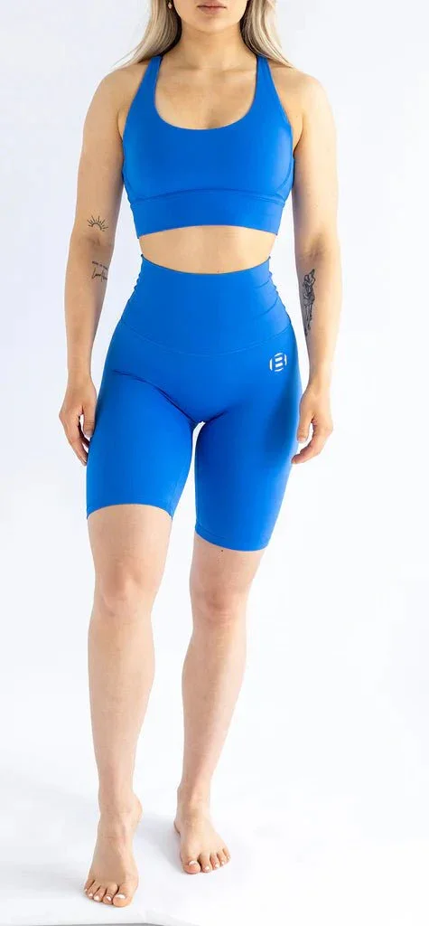 A woman in blue athletic wear, including a sports bra and high-waisted shorts, standing barefoot against a white background.