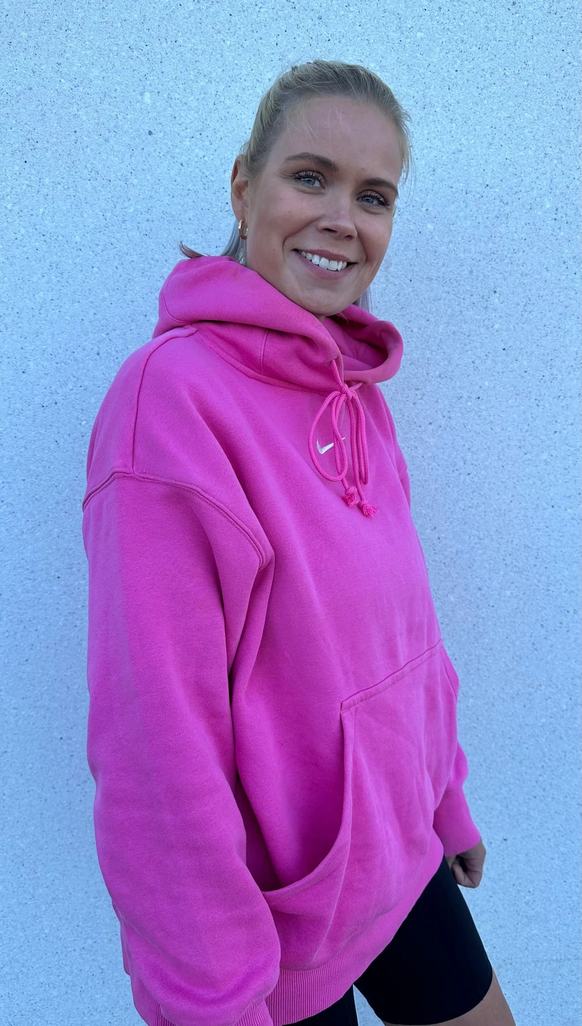 A woman with blonde hair tied back, wearing a bright pink Nike hoodie, smiling, standing against a light blue textured wall.