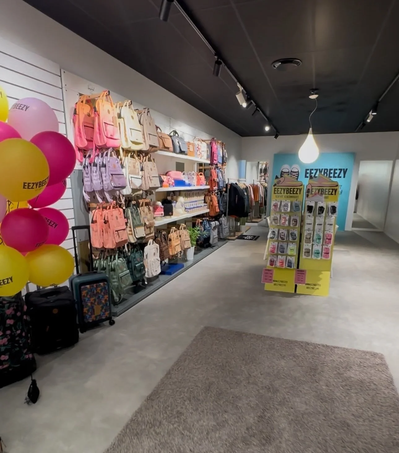 Inside a retail store display area with shelves of colorful backpacks, a stand with various small items, and balloons with the text 'EEZYBEEZY' on them. The store has a modern interior with a black ceiling and track lighting.