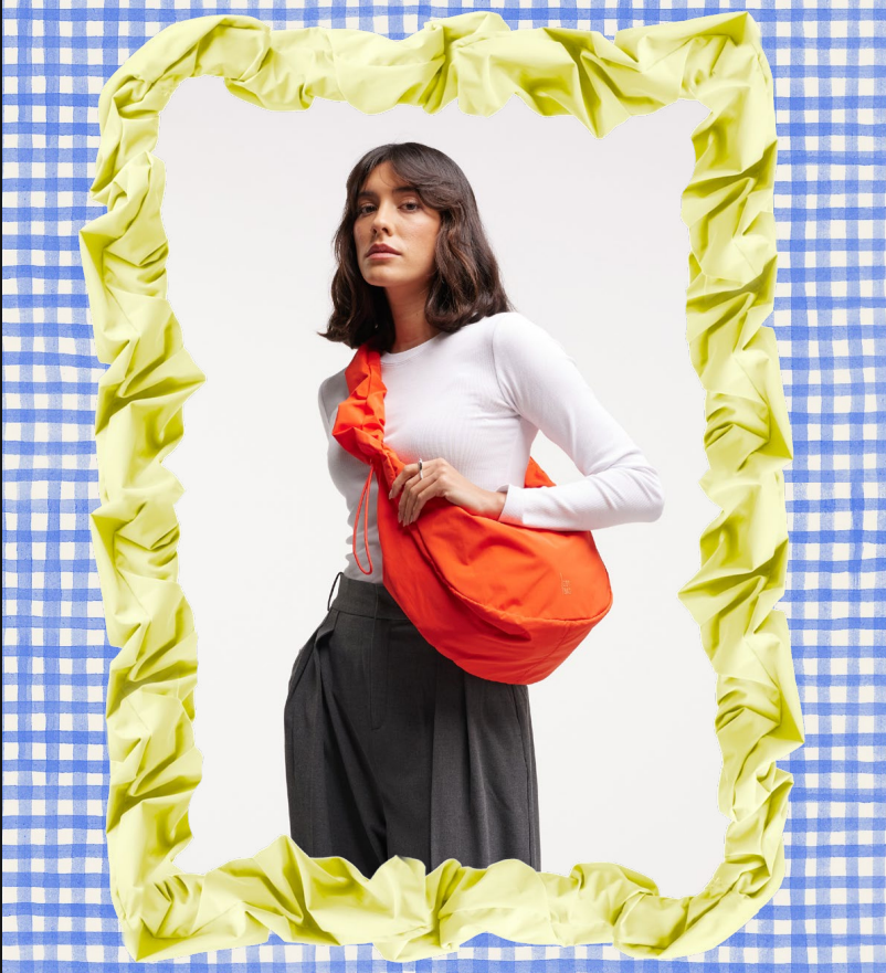 A woman with shoulder-length dark hair and a neutral expression, wearing a white long-sleeve top and dark gray pants, is standing against a plain background. She has an orange bag slung across her torso. The photo is framed with a crumpled yellow border and a blue-and-white checkered background.