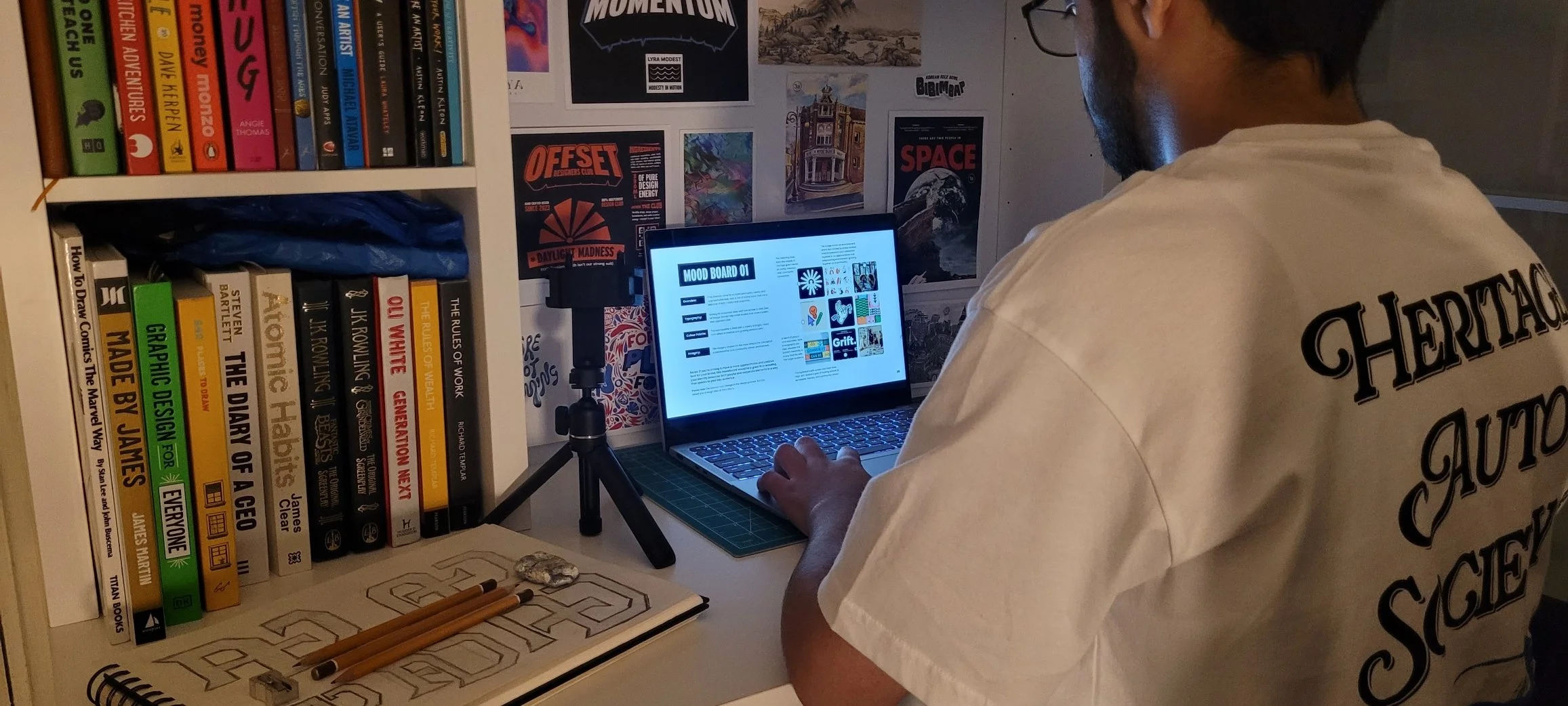 Person sitting at a desk using a laptop with a mood board on the screen. The desk has art supplies, a stone, and a notebook. Behind are bookshelves and wall posters, including space and art-themed images. The person wears a white T-shirt with black text on the back.