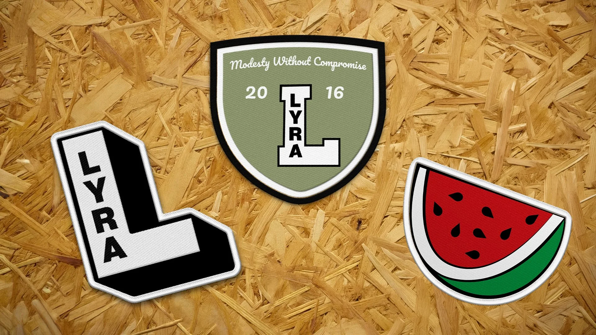 A patch with the text 'Modesty Without Compromise 2016' and the phrase 'LYRA' vertically in the center, mounted on a wooden surface with two additional patches: an L-shaped patch with the word 'LYRA' and a watermelon patch depicting a slice of waterm