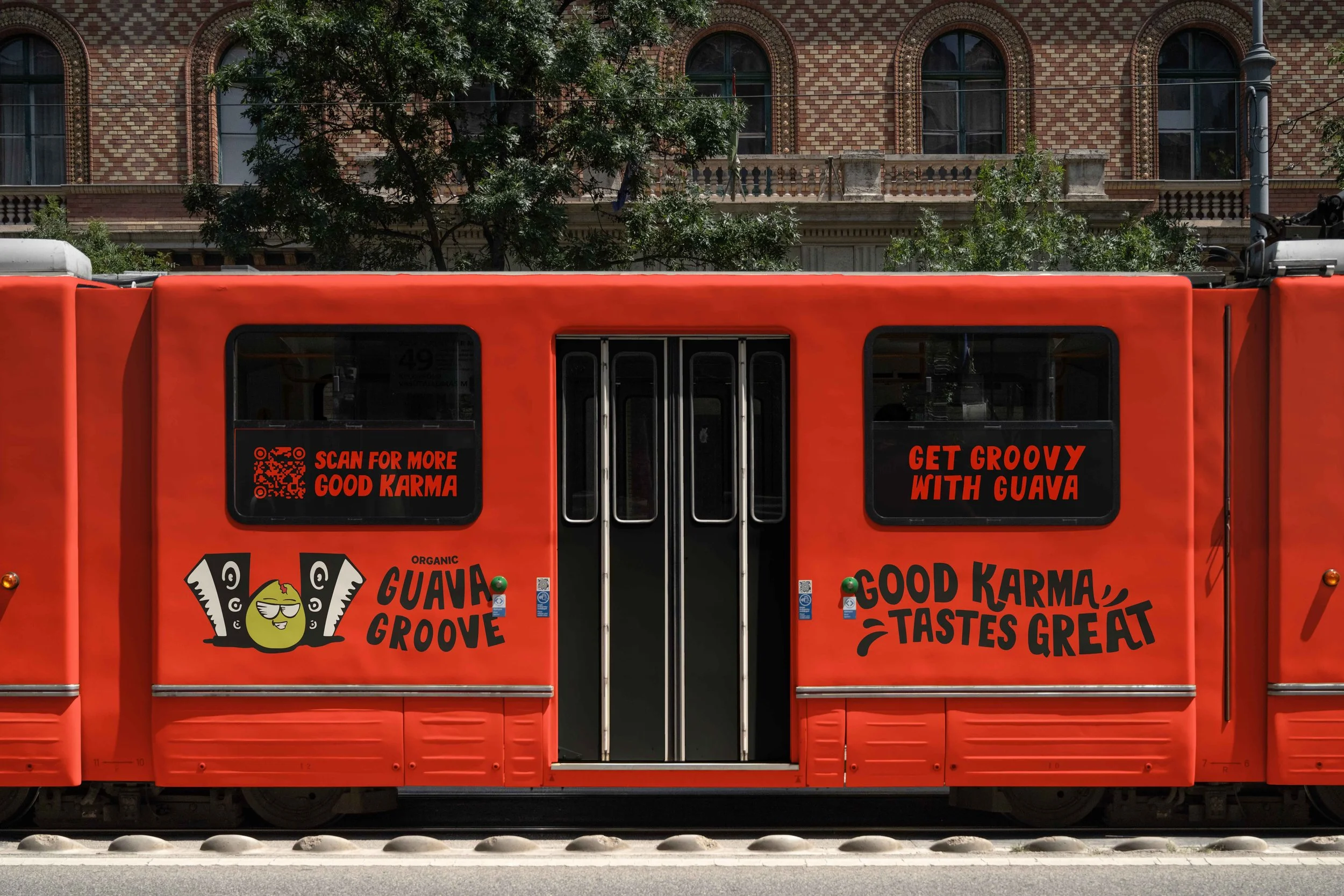 A bright orange tram with promotional advertisements for Guava Groove, featuring a cartoon guava character and the phrases 'Scan for more good karma' and 'Get groovy with guava.' The background includes a brick building with arched windows and greene