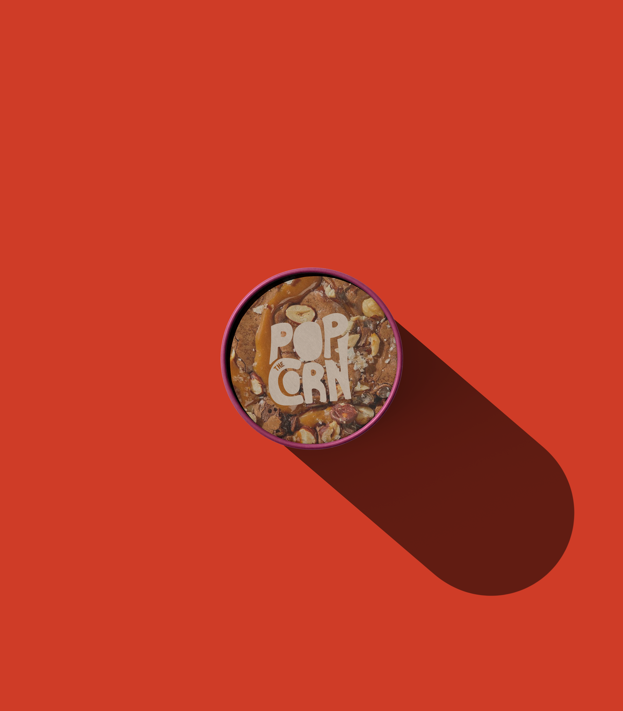 A round container of popcorn with a pink rim, labeled as "Pop the corn" on top, on a solid red background with a shadow extending to the right. designed by Abubakr Moolla Brand Designer from East London.