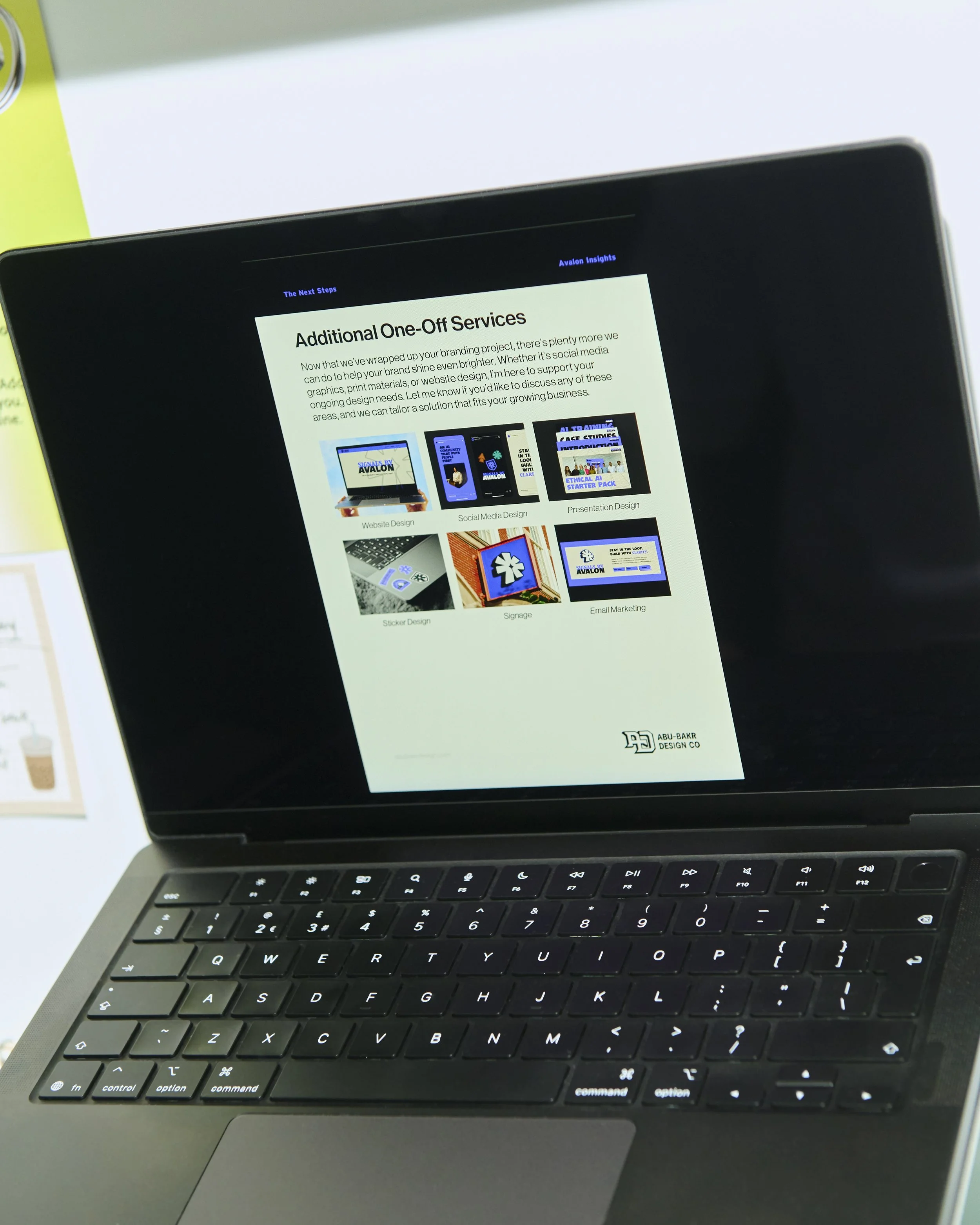 Close-up of a laptop with a black keyboard and a screen displaying a webpage titled 'Additional One-Off Services.' The webpage contains text and images related to branding, website, social media, presentation design, sticker design, signage, and email marketing services.