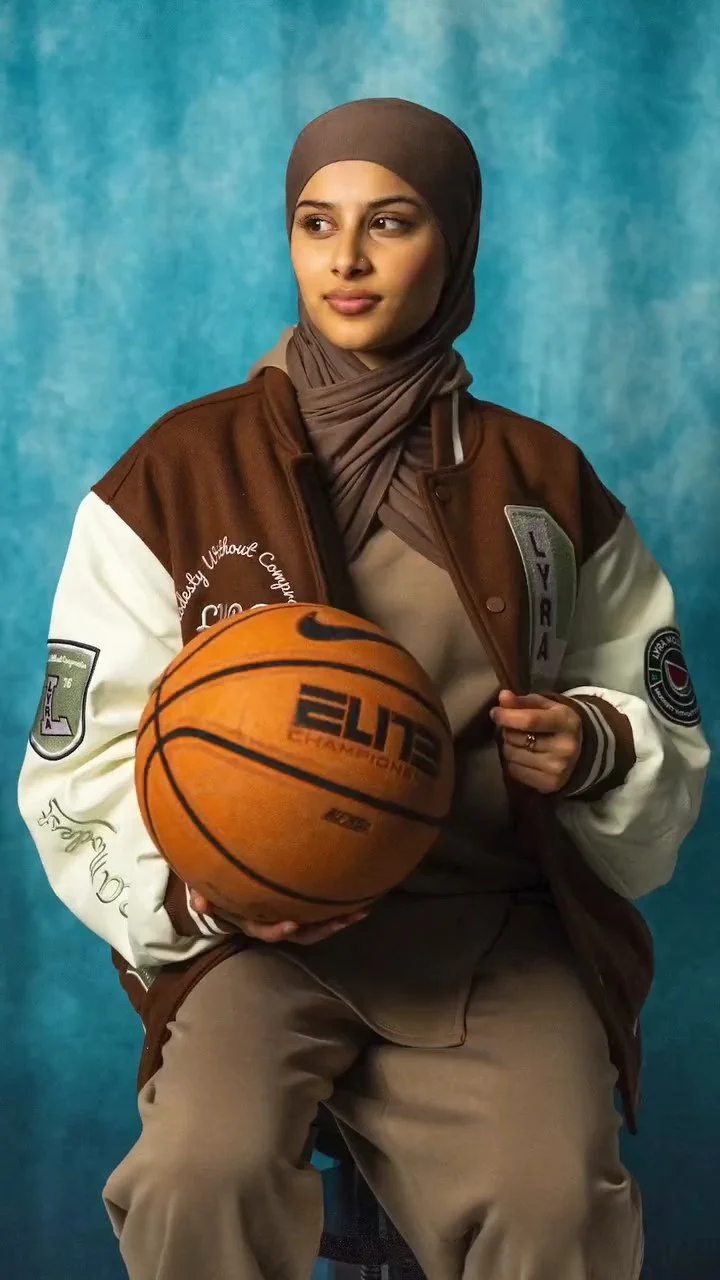 A young woman wearing a brown and white letterman jacket and a brown hijab, sitting and holding a basketball, with a blue textured background.