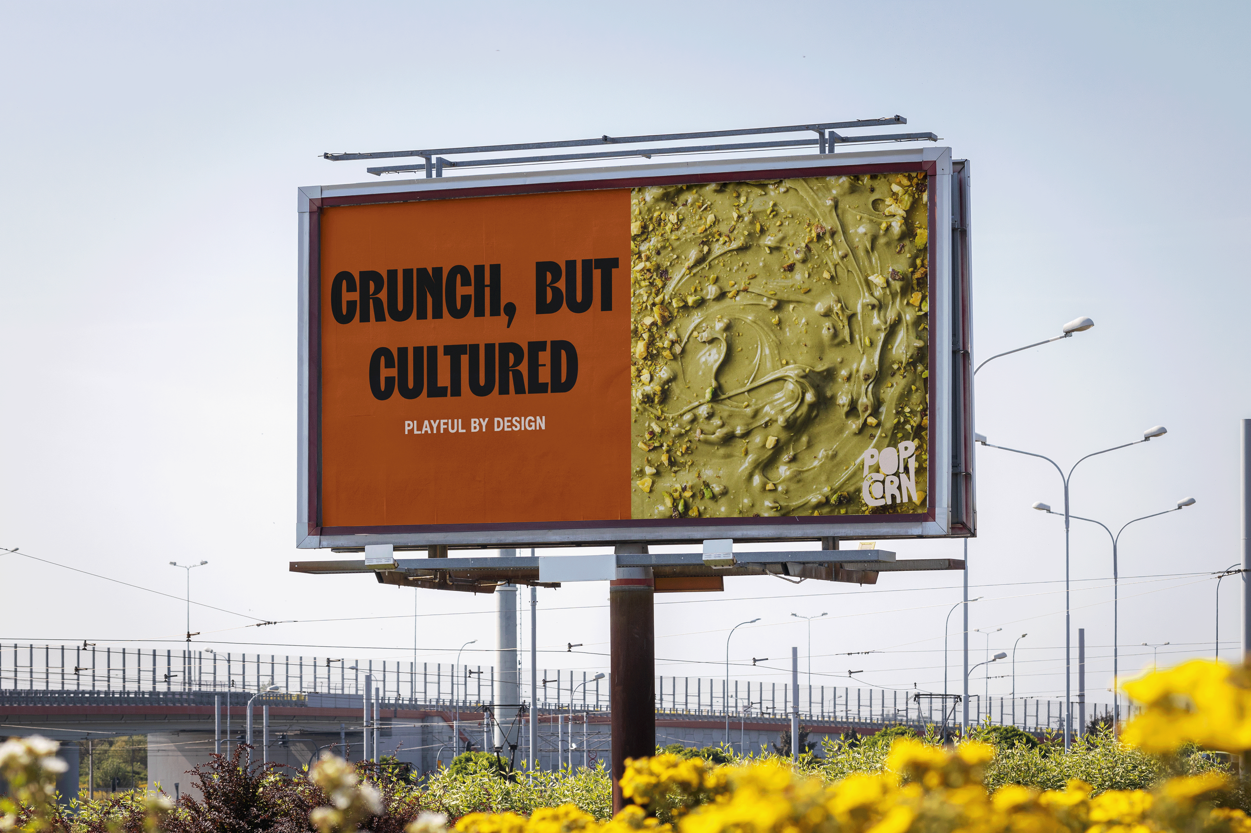 Billboard advertising a snack with the words 'Crunch, but cultured, playful by design' and an image of green swirled snack with chopped nuts, with a background of streetlights and an elevated railway. designed by Abubakr Moolla Brand Designer from Ea