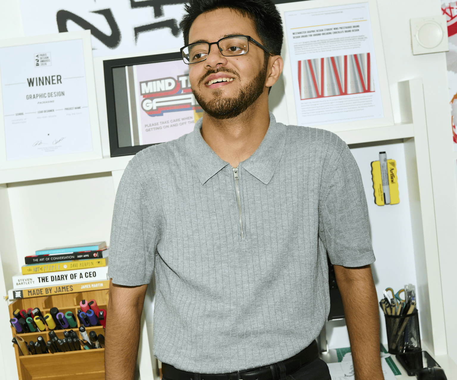 A young man with glasses and a beard smiling in an office or classroom setting, standing in front of shelves with books, certificates, and art supplies.