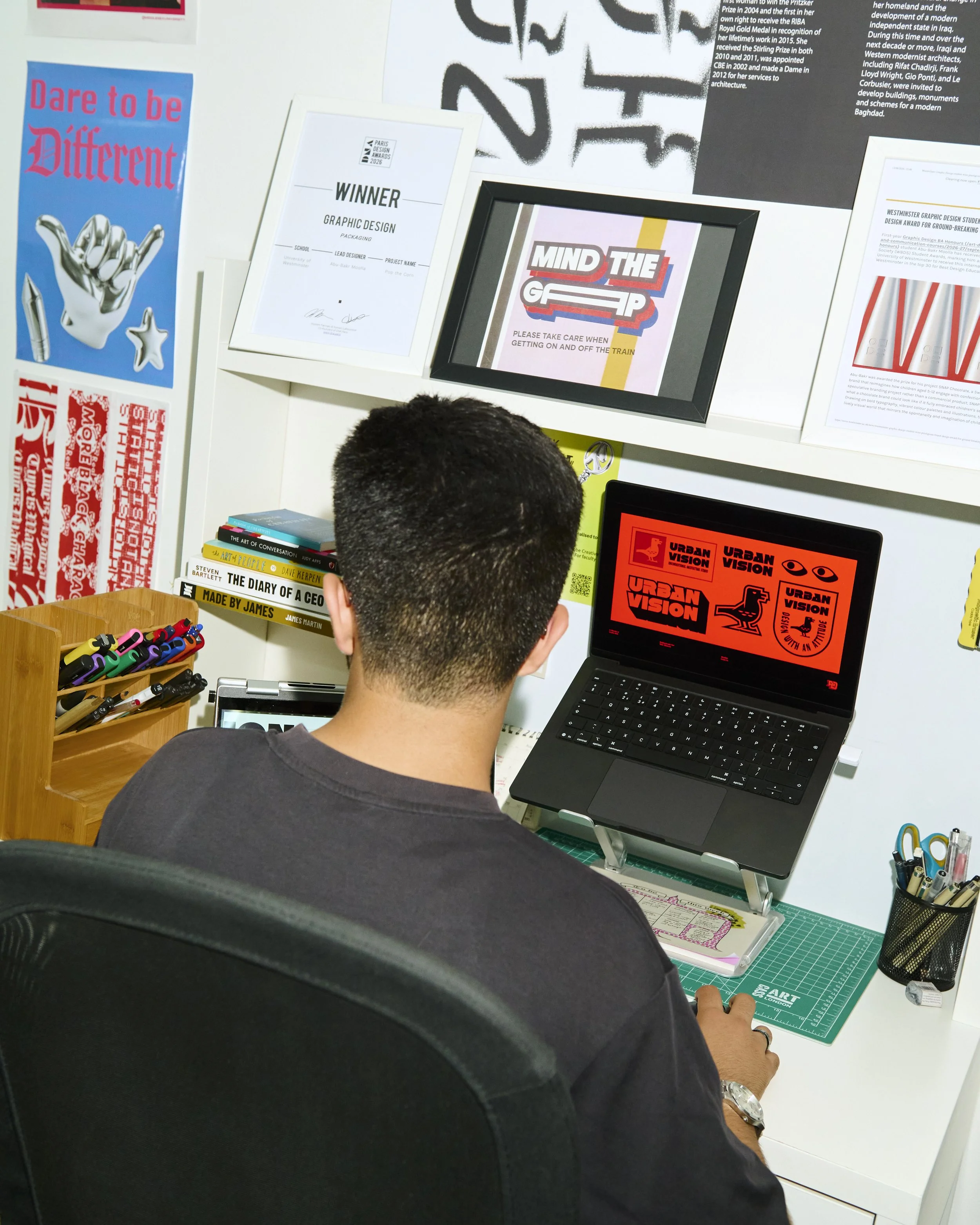 Person working at a desk with a laptop, surrounded by posters, certificates, and office supplies in a creative workspace.