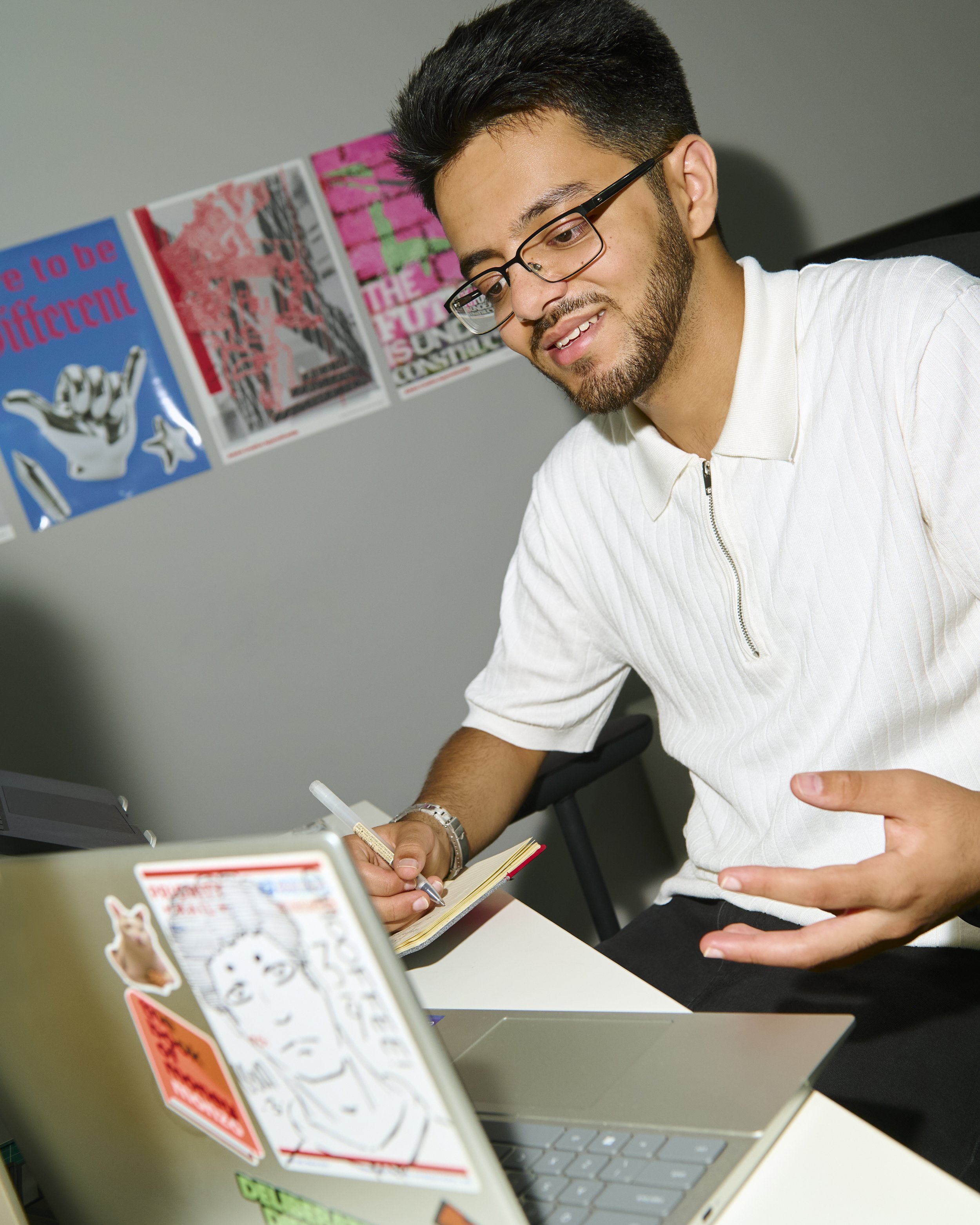A man with dark hair, glasses, and a beard is smiling and writing in a notebook while seated at a table with a laptop. In the background, there are colorful posters on the wall.