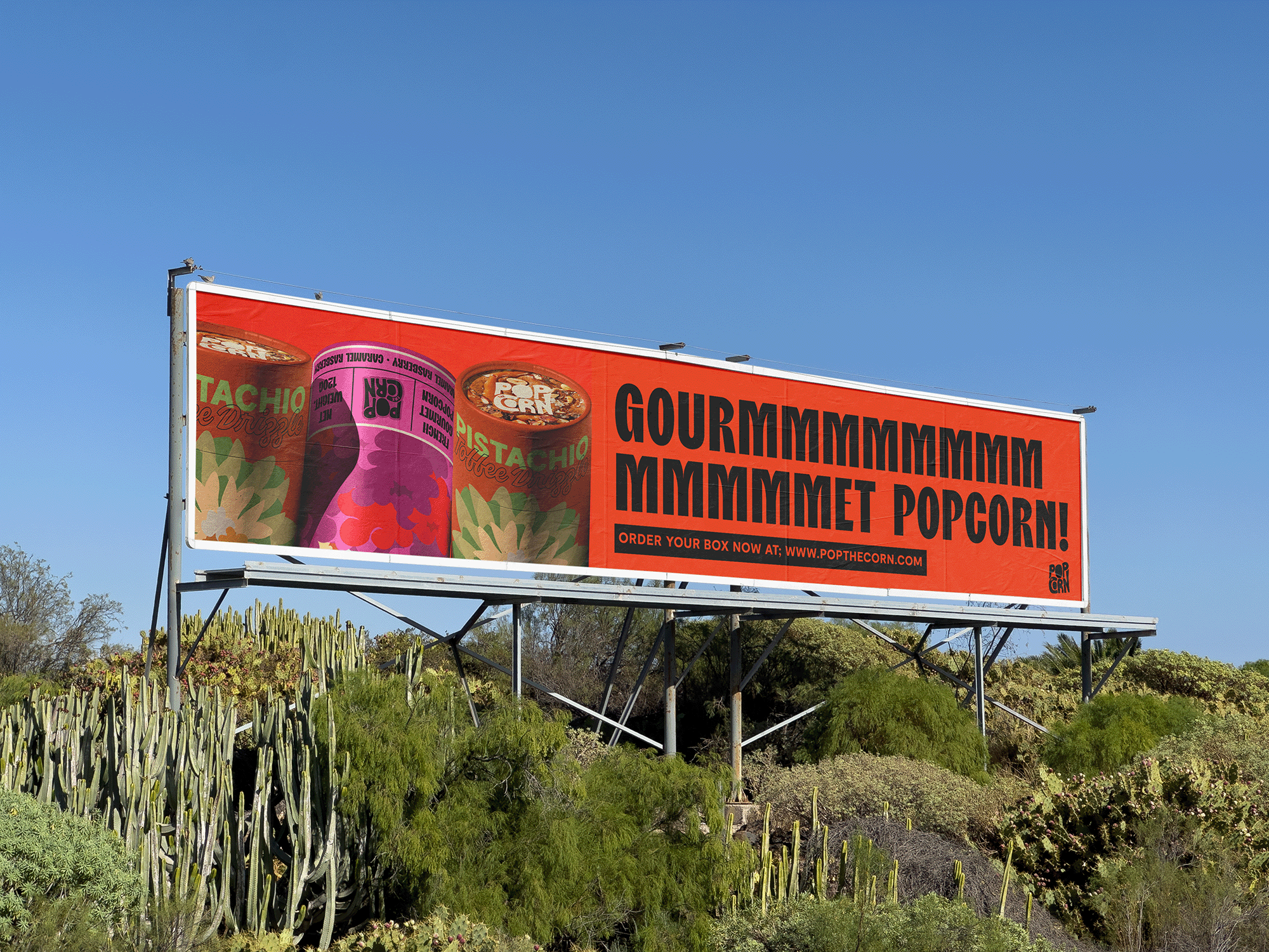 Billboard advertising popcorn from Pop the Corn, featuring images of colorful popcorn boxes and a bowl of popcorn, with the text 'GOURMMGGMMM MMMMMMPOP!' and a website link.