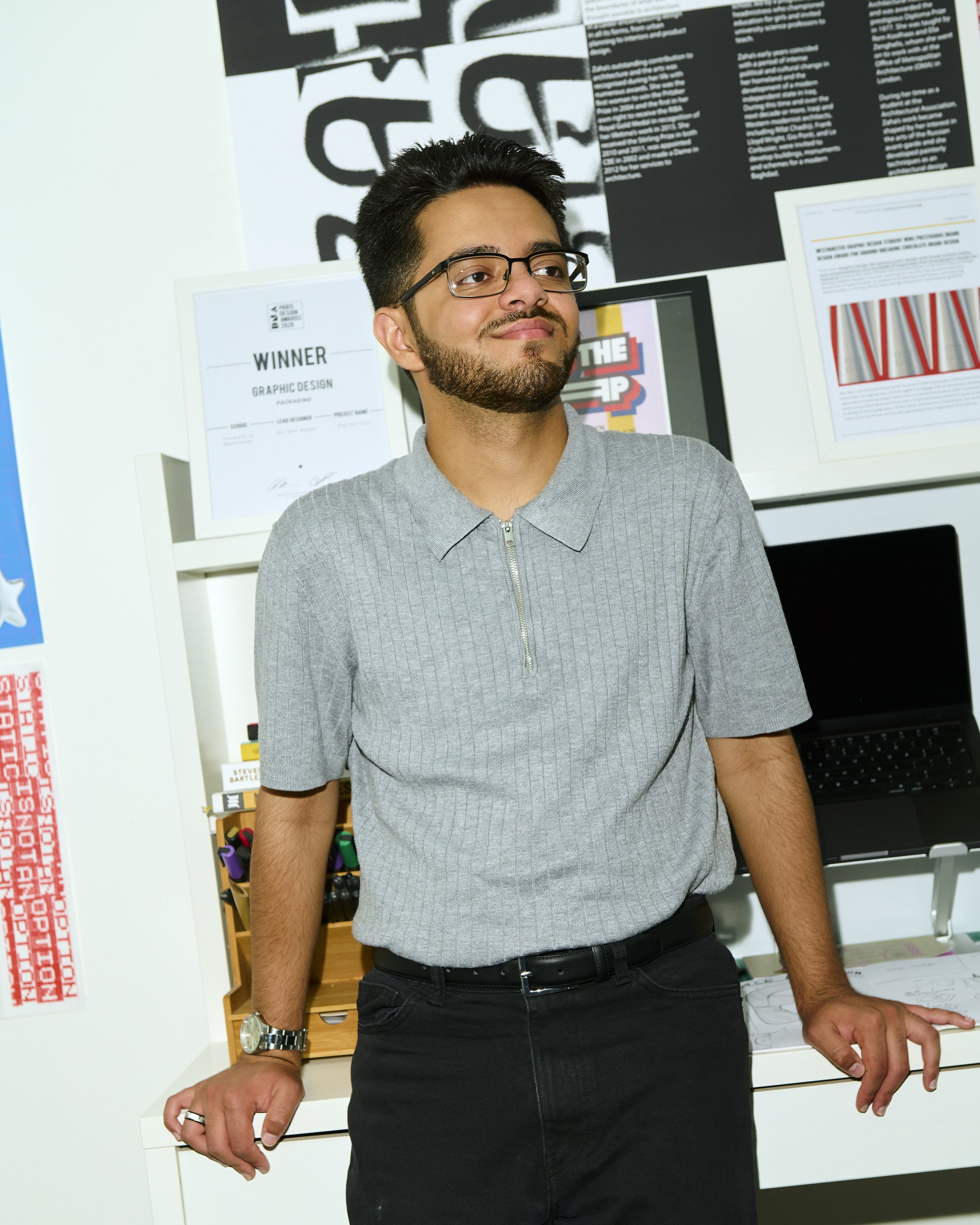 A young man with black hair, glasses, and a beard wearing a gray zip-up shirt. He is standing in front of a wall decorated with various posters and artwork, including a certificate and framed print, leaning slightly on a white desk or table with his right hand resting on it.