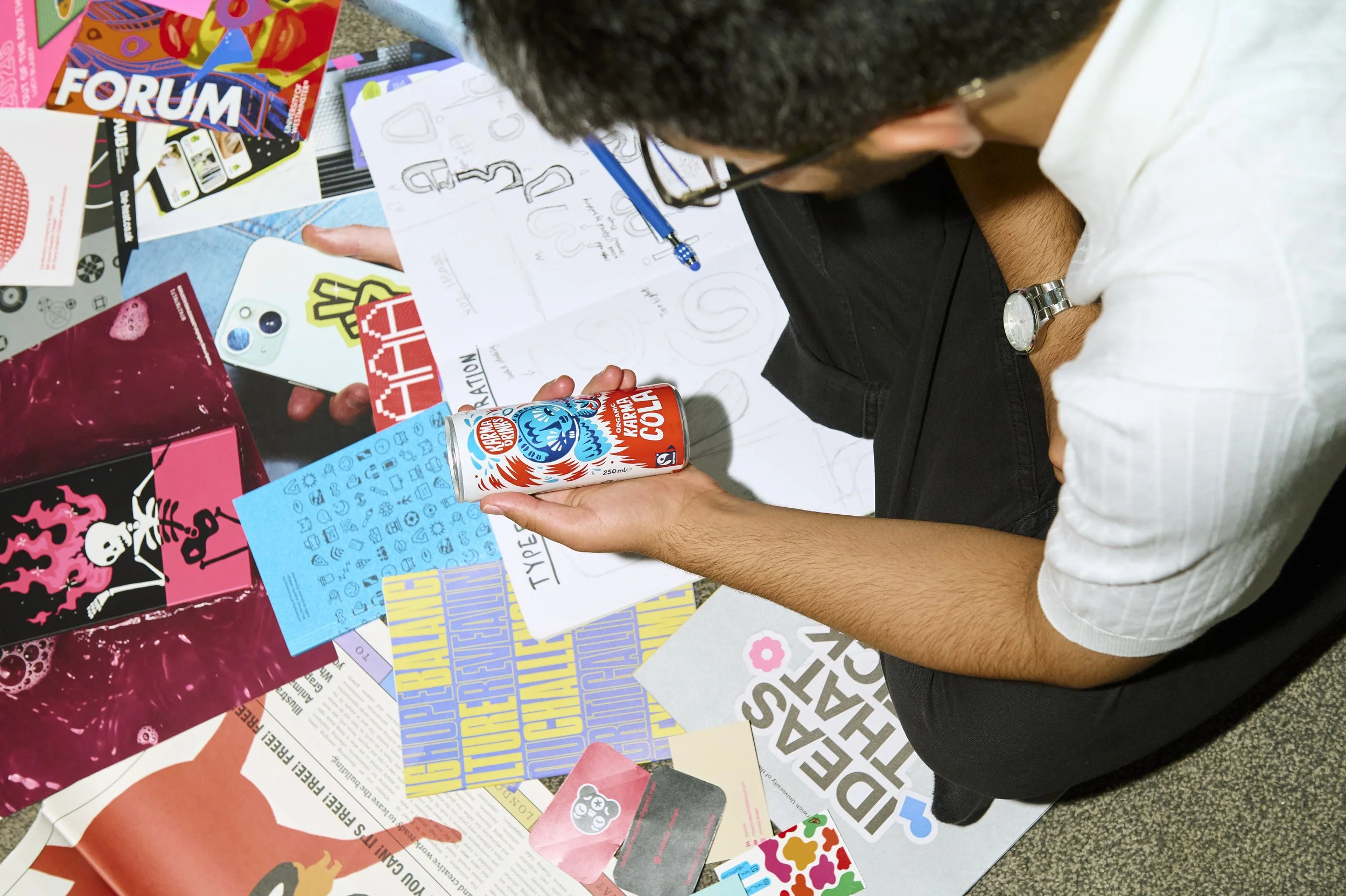 A person with glasses and a watch is sitting at a table surrounded by various colorful papers, stickers, and flyers, drawing or writing on a sheet of paper.