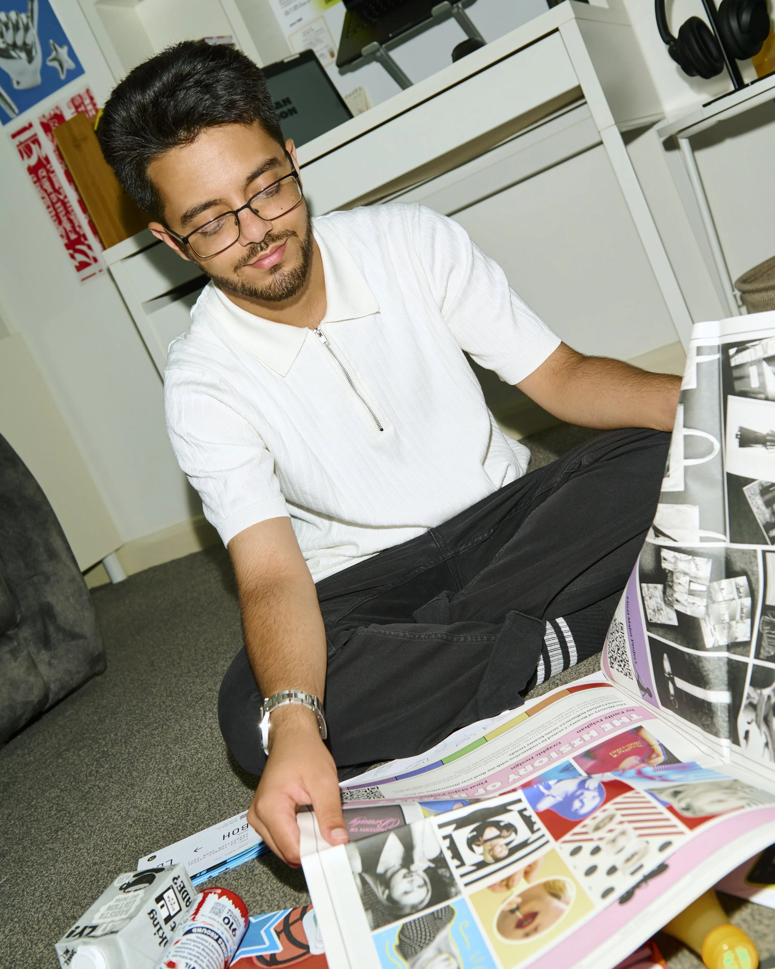 A young man with glasses and beard, wearing a white zip-up shirt, sitting on the floor, flipping through a colorful magazine or catalog with comic and pop culture images.