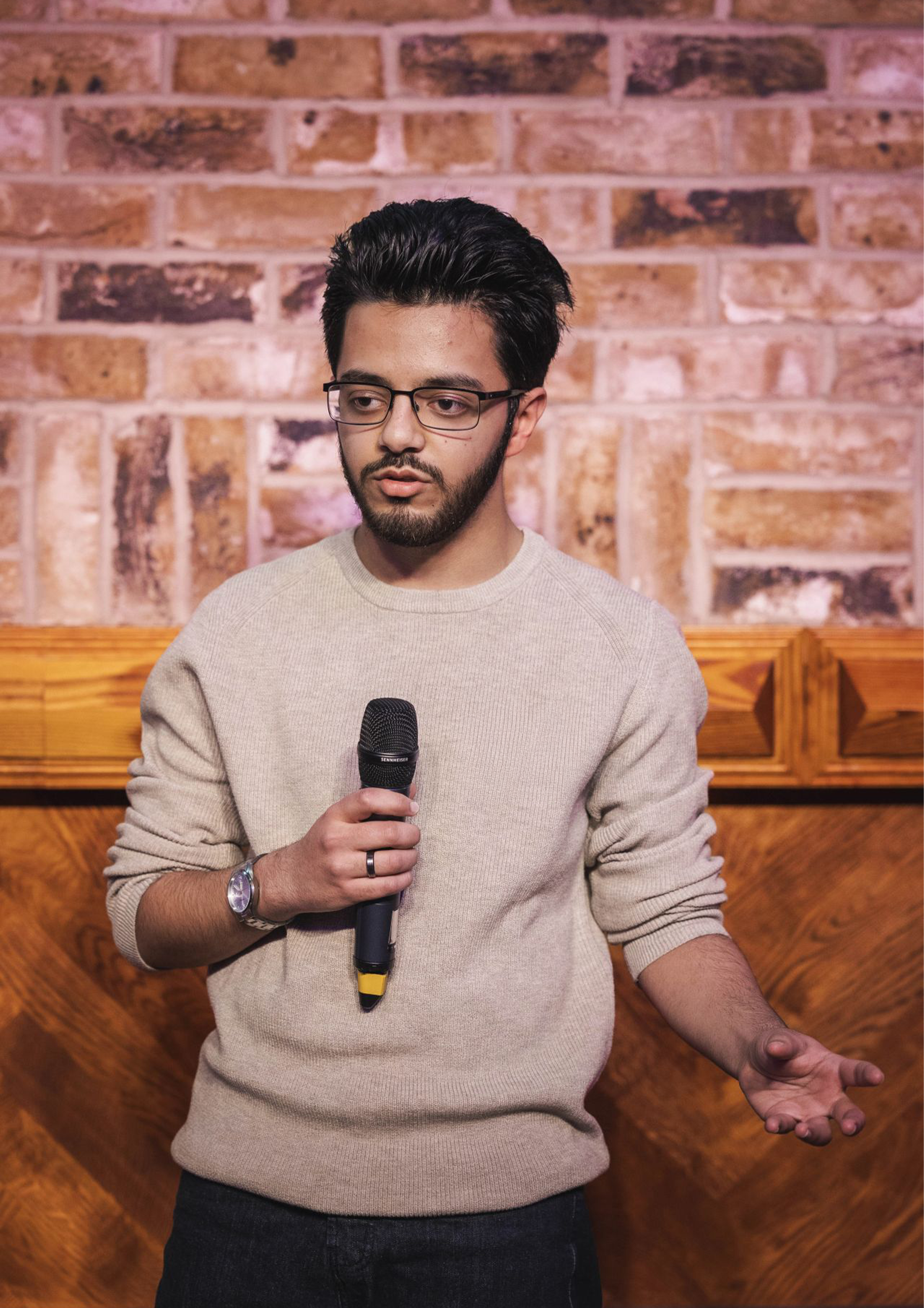 A young man with dark hair, glasses, and a beard, holding a microphone in his right hand and gesturing with his left hand, standing in front of a brick wall background.