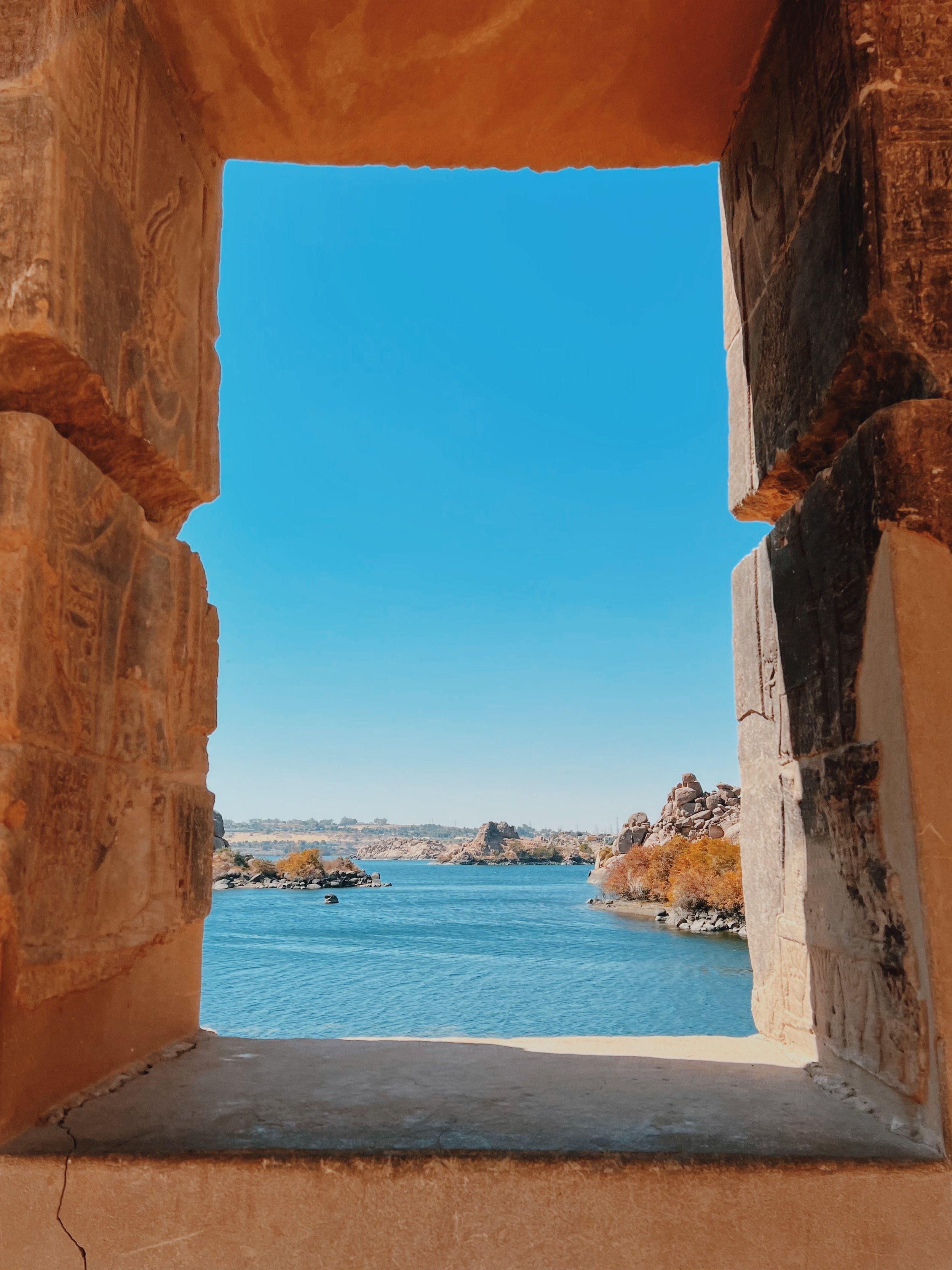 An ancient stone window at Philae Temple framing the blue Nile, granite islands and autumn trees beyond