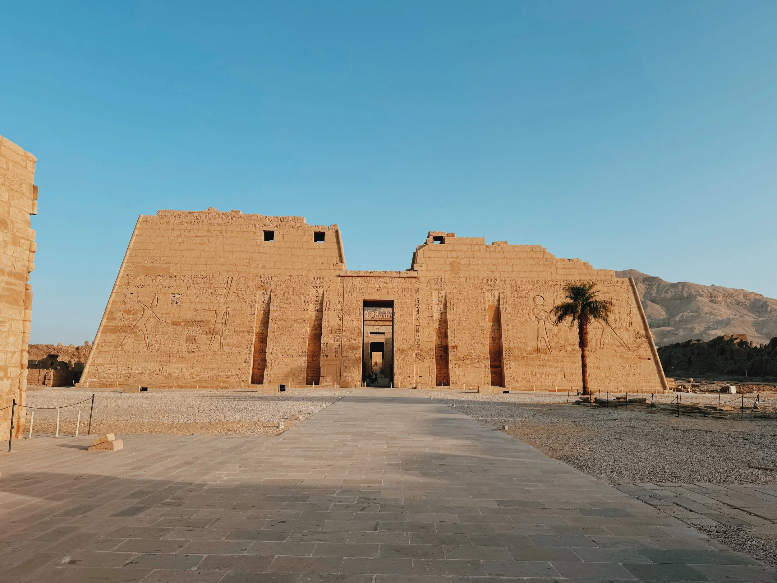 The towering carved first pylon of Medinet Habu temple on Luxor's West Bank, a stone path leading to its doorway and the Theban hills beyond