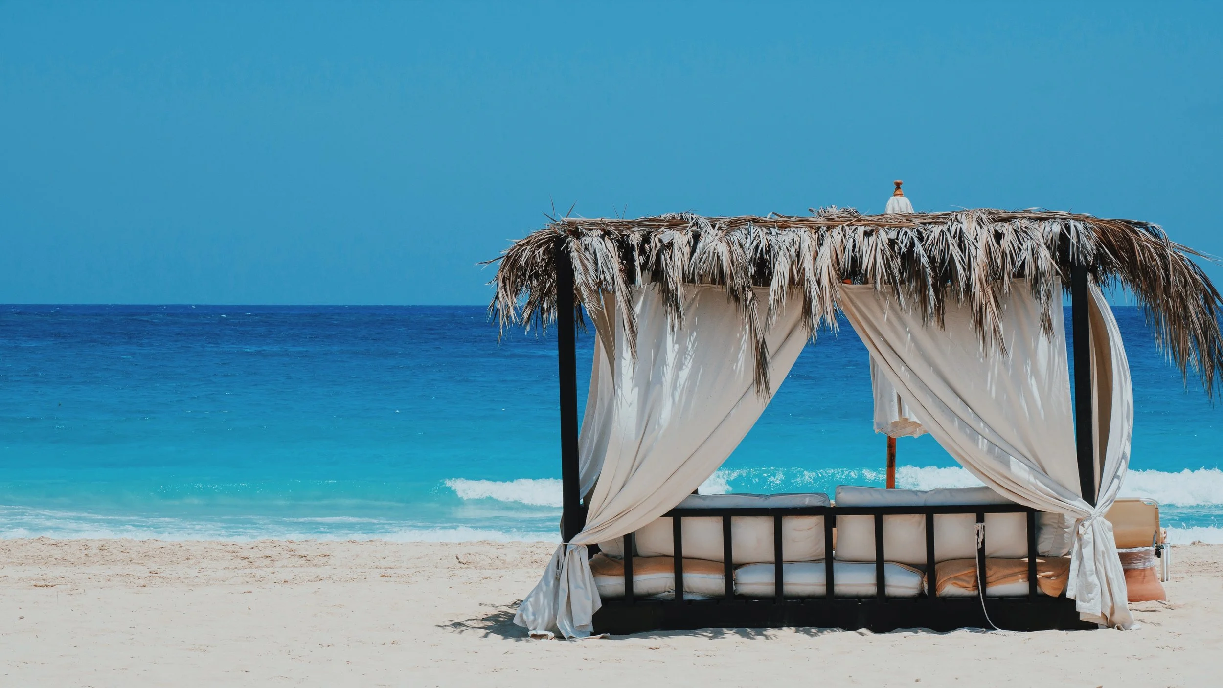 A palm-thatched cabana with white curtains on the beach at Sidi Heneish, facing the turquoise Mediterranean.