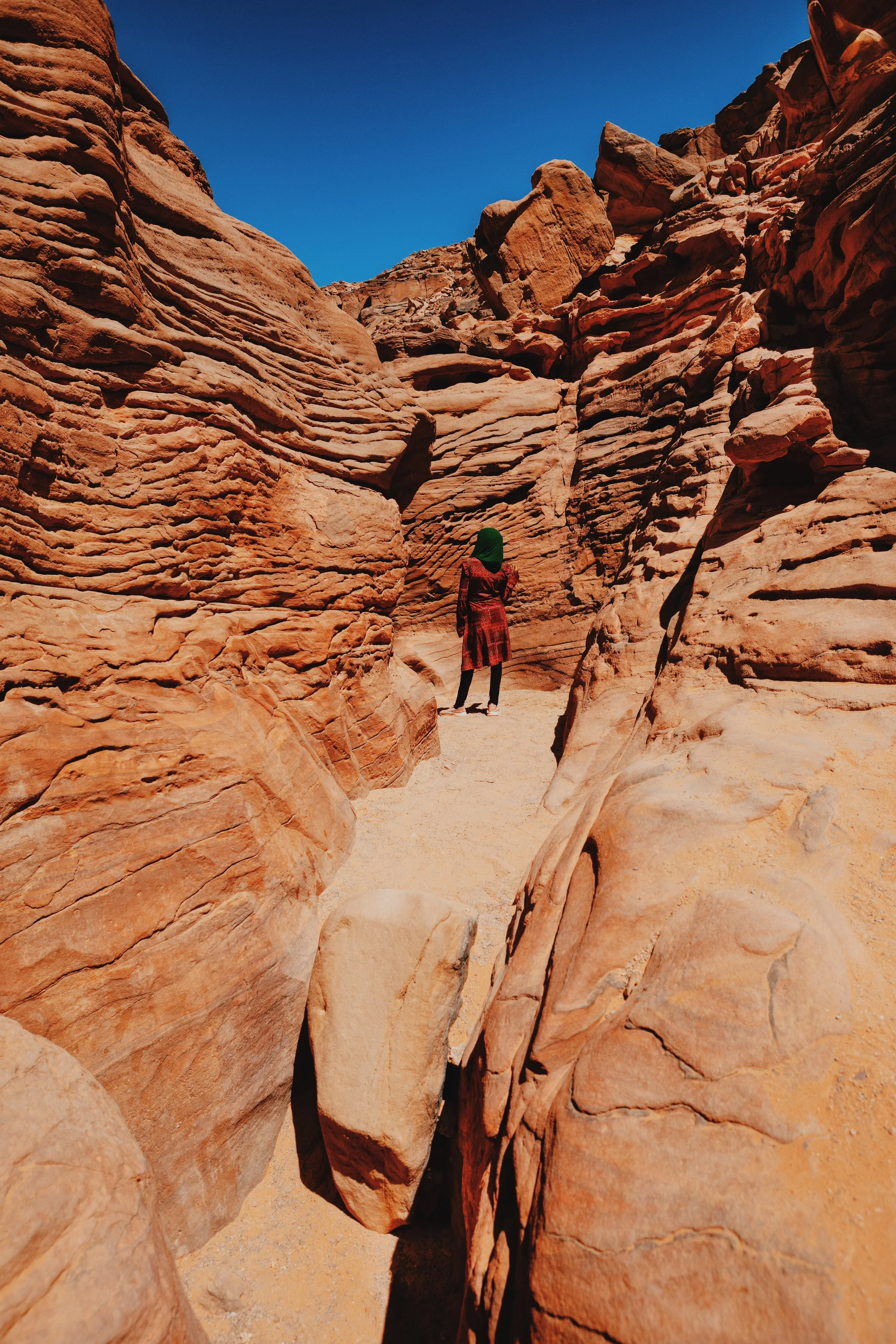 A woman in a green headscarf walking between the swirling sandstone walls of the Colored Canyon in South Sinai.