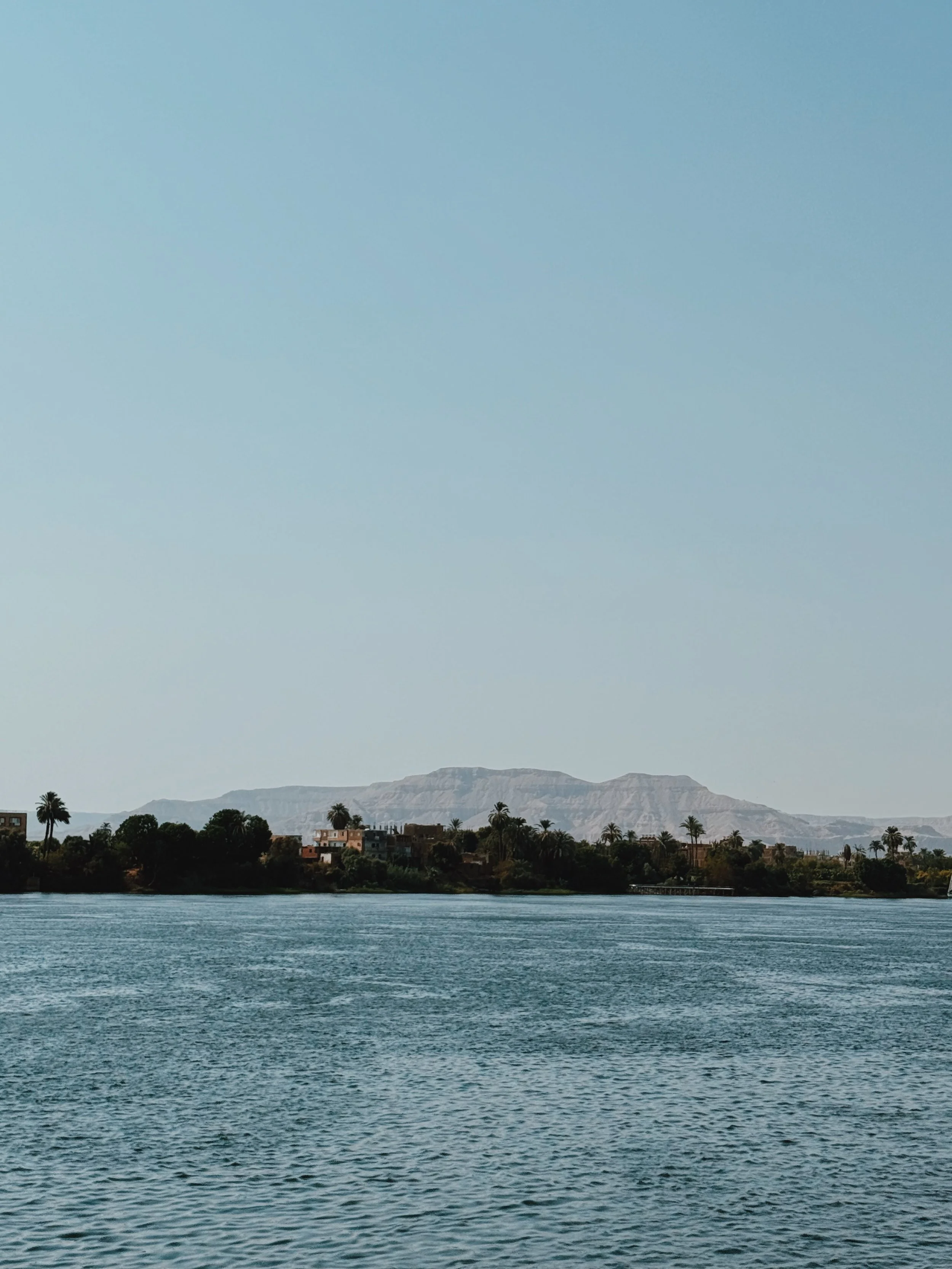 The Nile at Luxor looking across to the palm-lined West Bank and the Theban hills under a clear sky