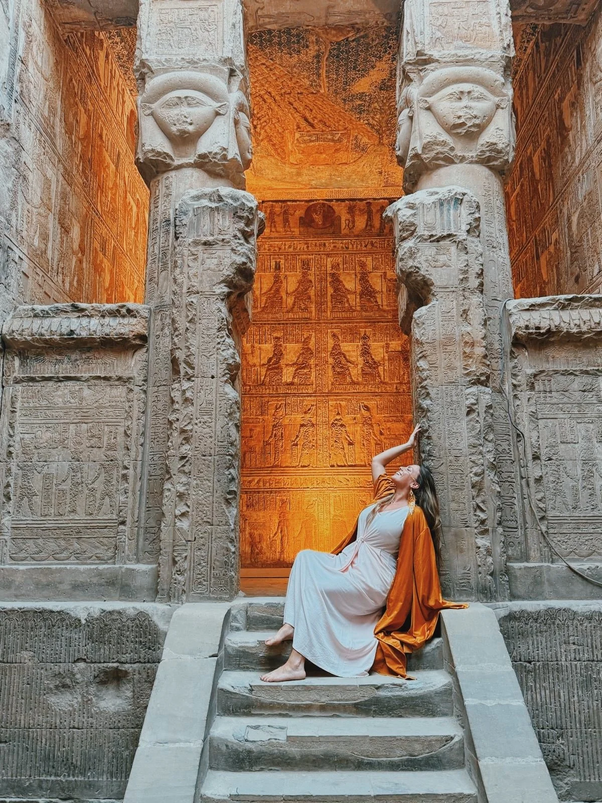 A traveler in a white dress and saffron robe raising her hand in joy on the steps between the Hathor columns at Dendera.