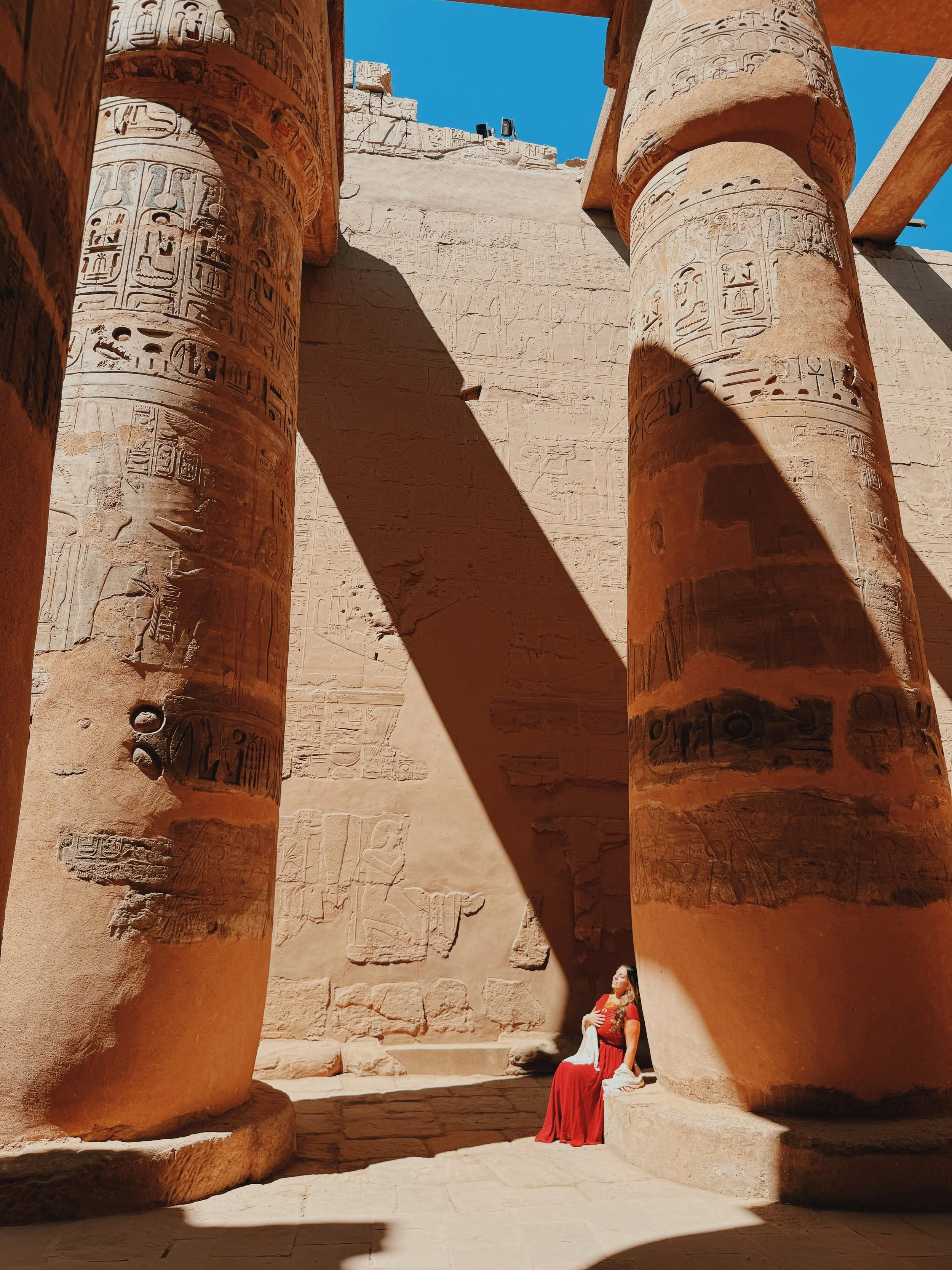 Woman in a red dress seated in sunlight at the base of the giant carved columns in the Hypostyle Hall of Karnak Temple, Luxor