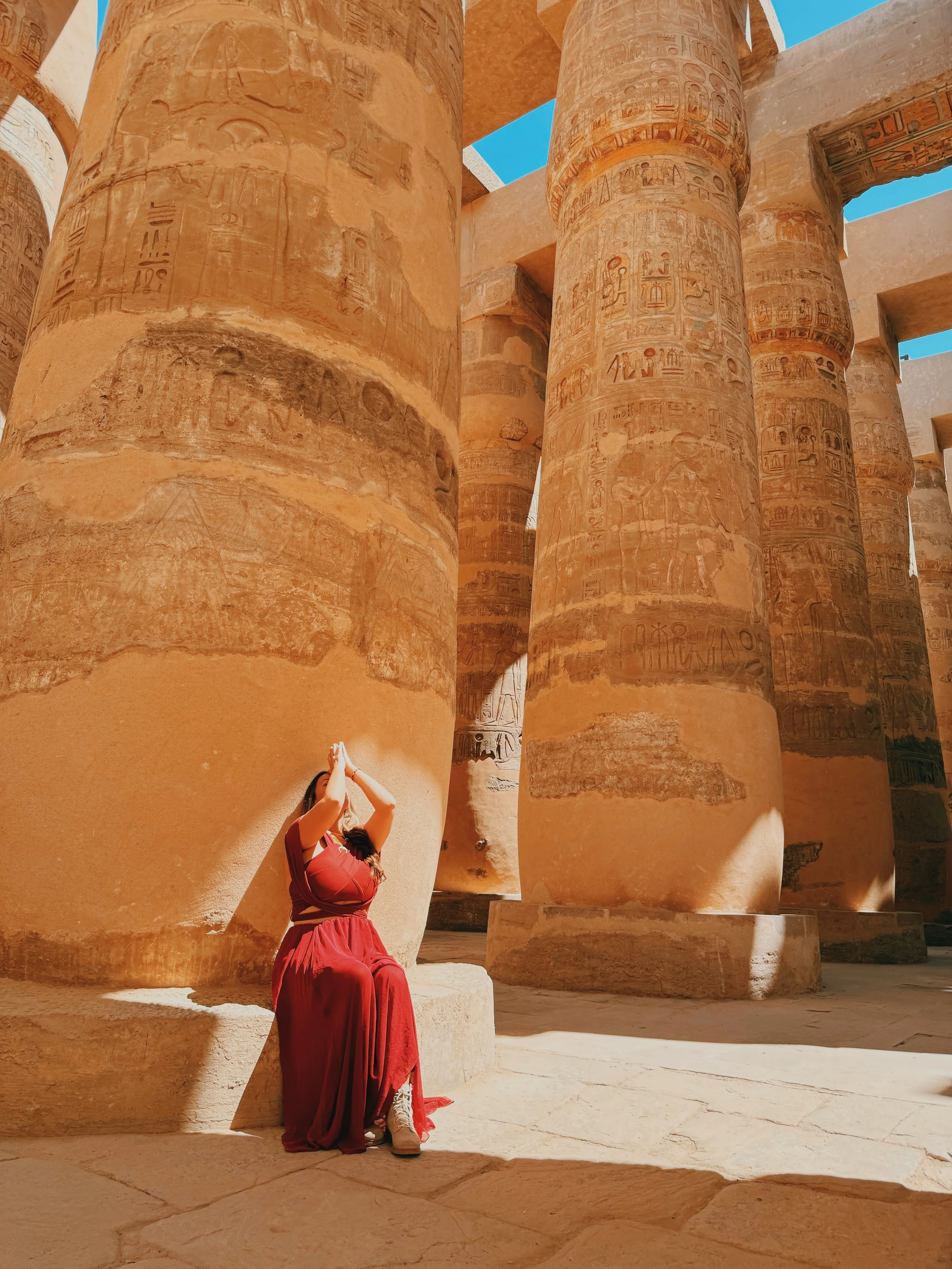 Woman in a red dress seated beneath the giant columns of the Hypostyle Hall at Karnak Temple, Luxor, face and hands raised to the sun