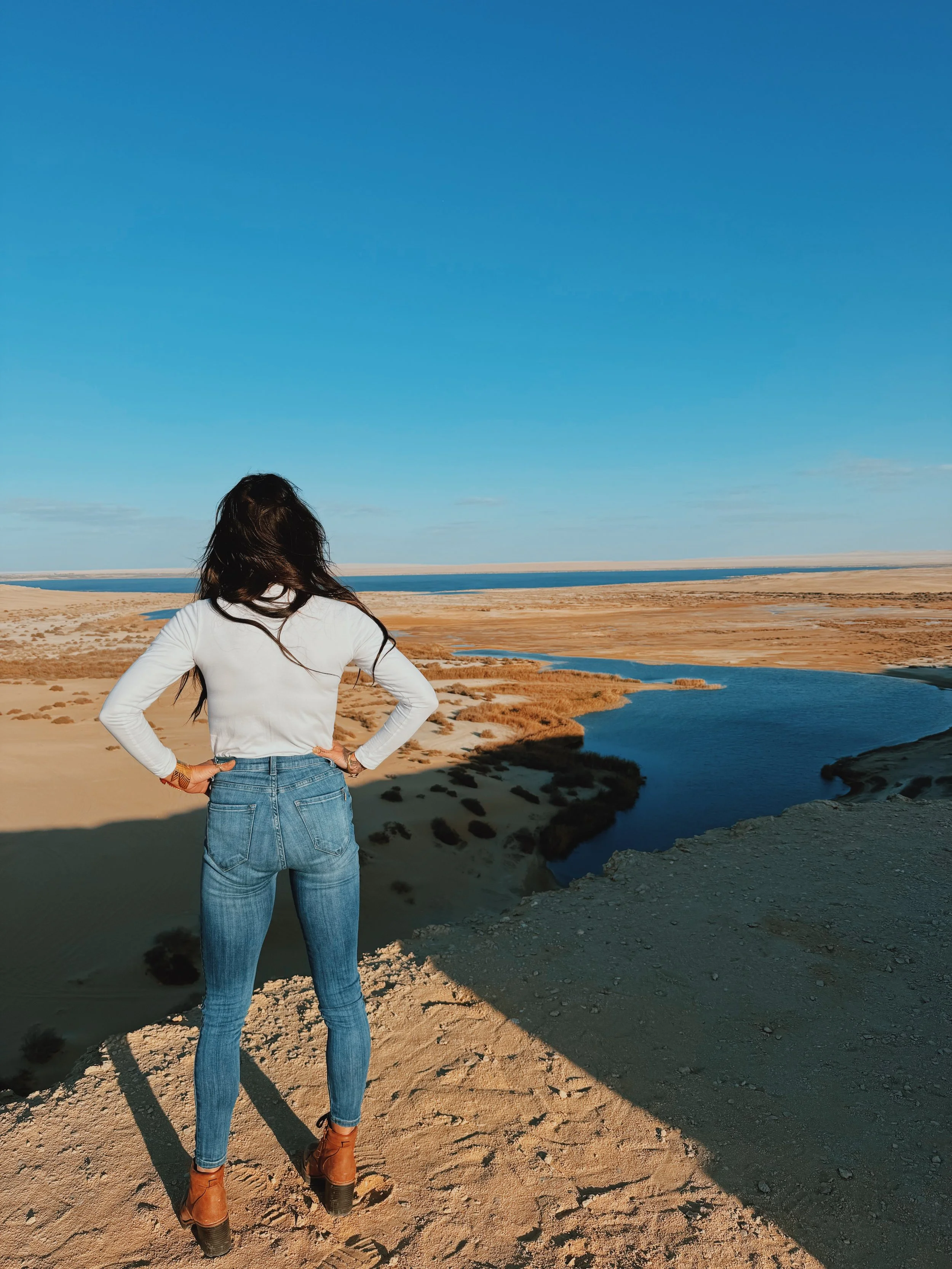 A woman in a white jacket standing at the water’s edge in the open Egyptian desert.