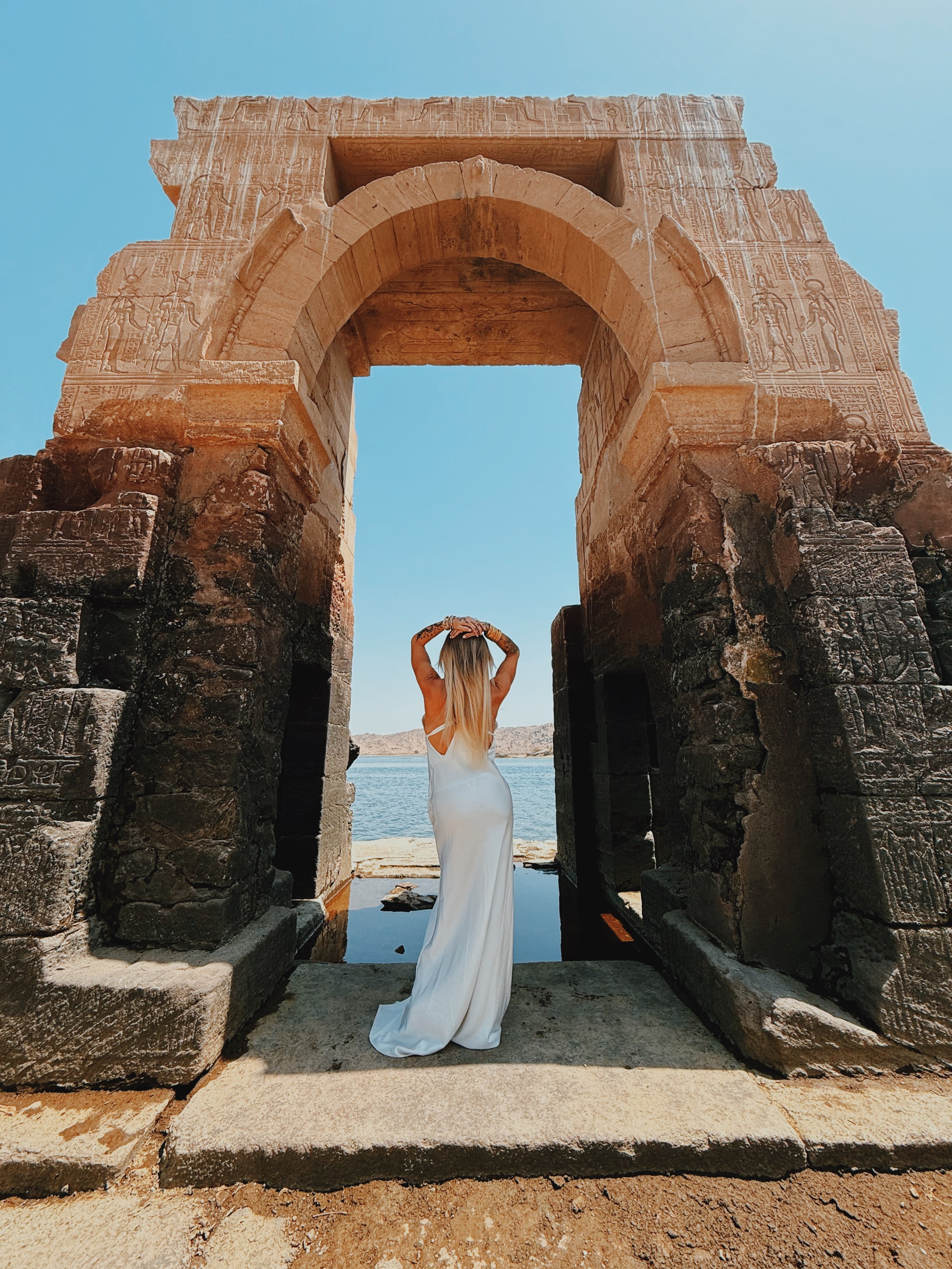 Woman in a white dress beneath an ancient carved stone arch at Old Philae Site, Aswan, looking out over the Nile where guests enter the water