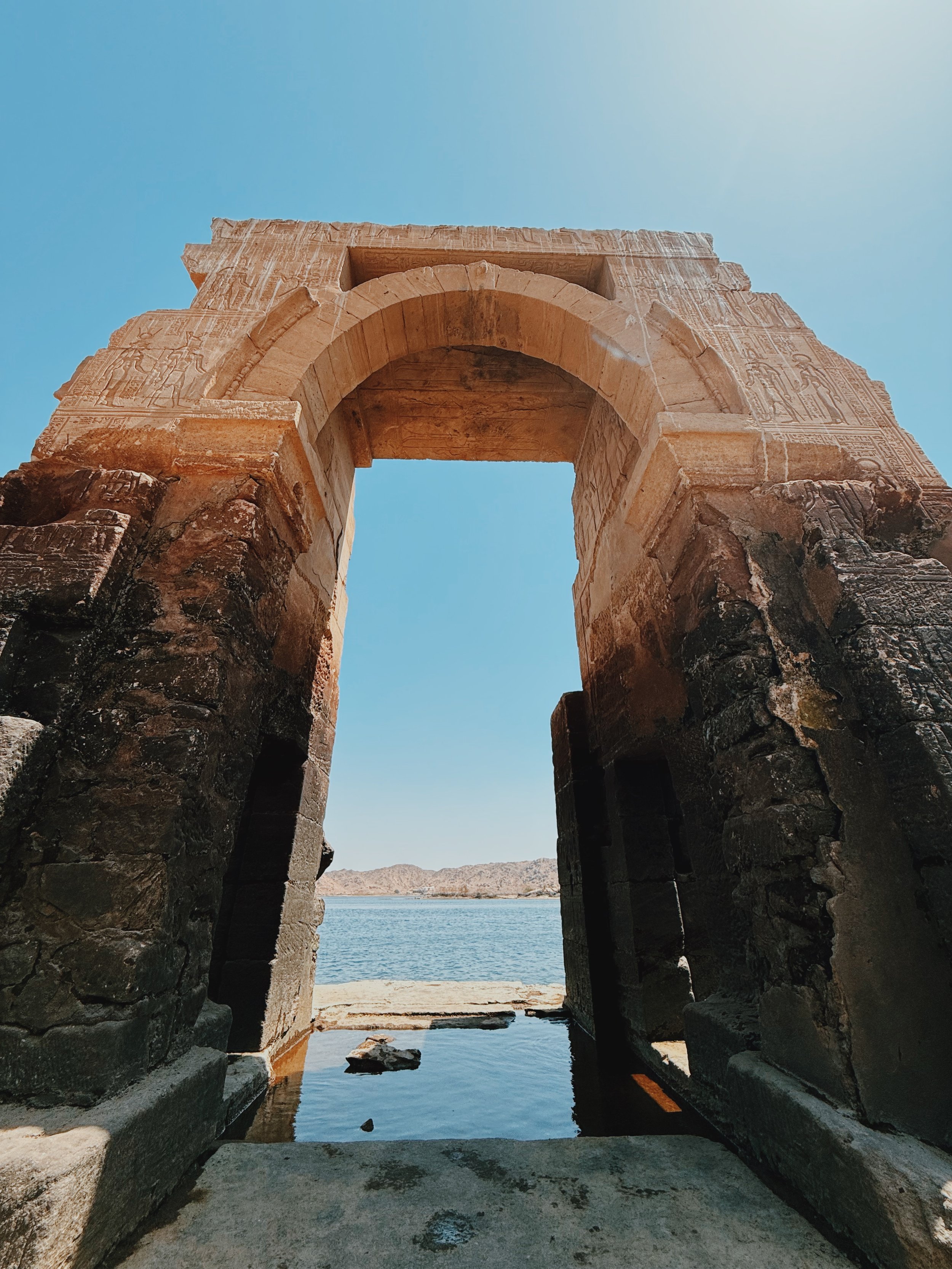 An ancient carved stone archway at old Philae near Aswan, framing the Nile and the desert hills beyond.