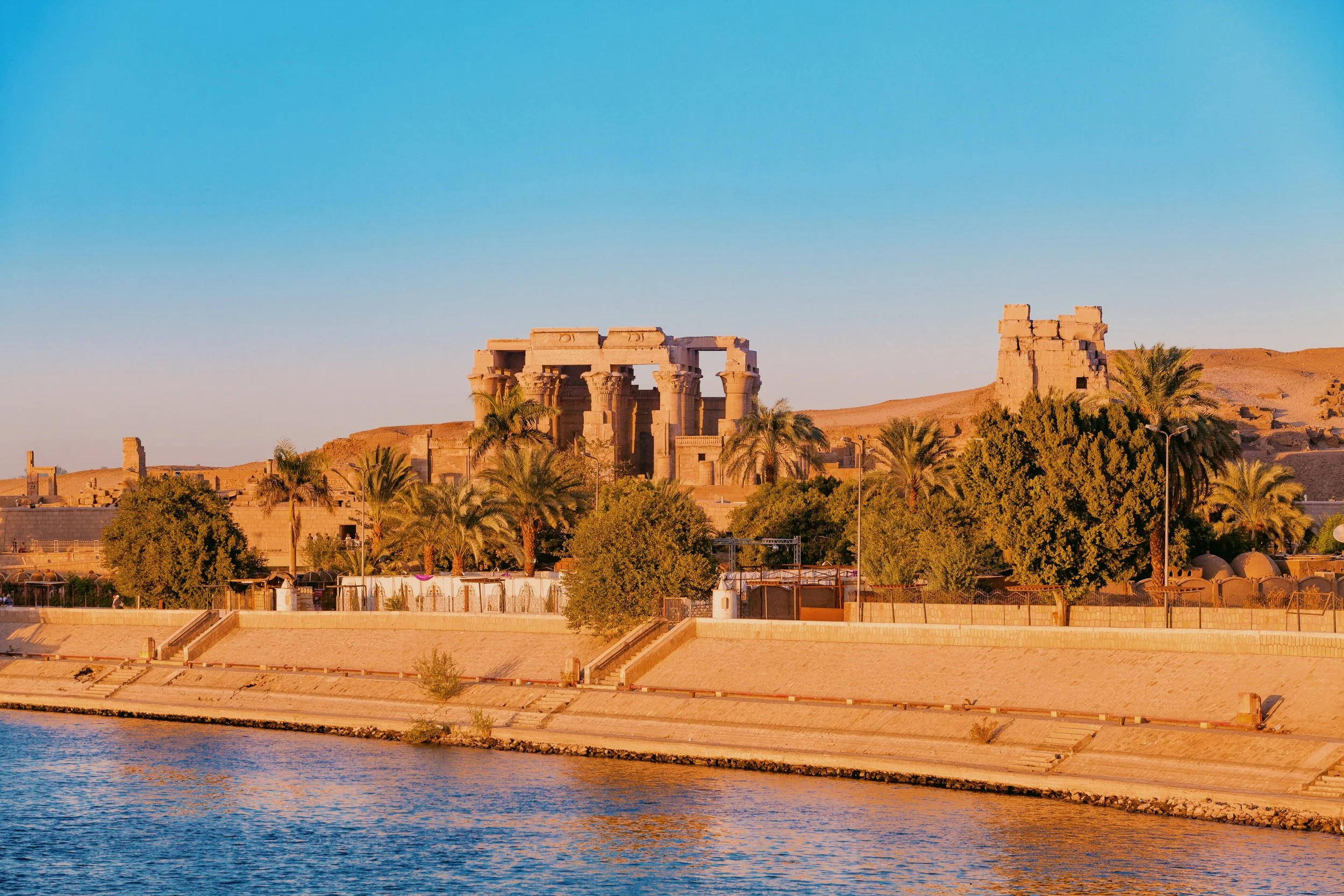 The Temple of Kom Ombo rising above palm trees on the bank of the Nile in golden afternoon light.