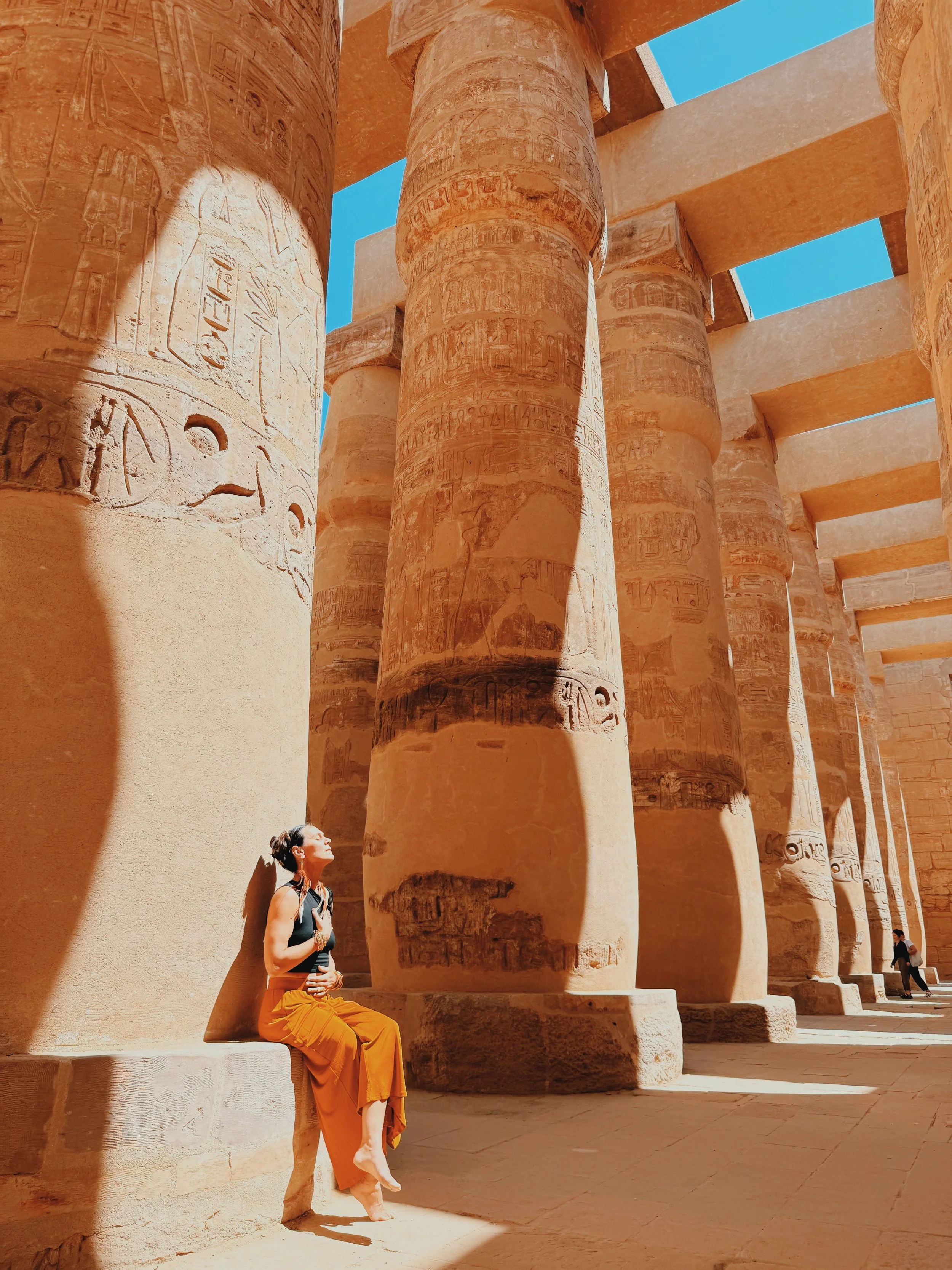 Woman in an orange skirt seated against a giant column in the Hypostyle Hall of Karnak Temple, Luxor, eyes closed and face turned to the sun