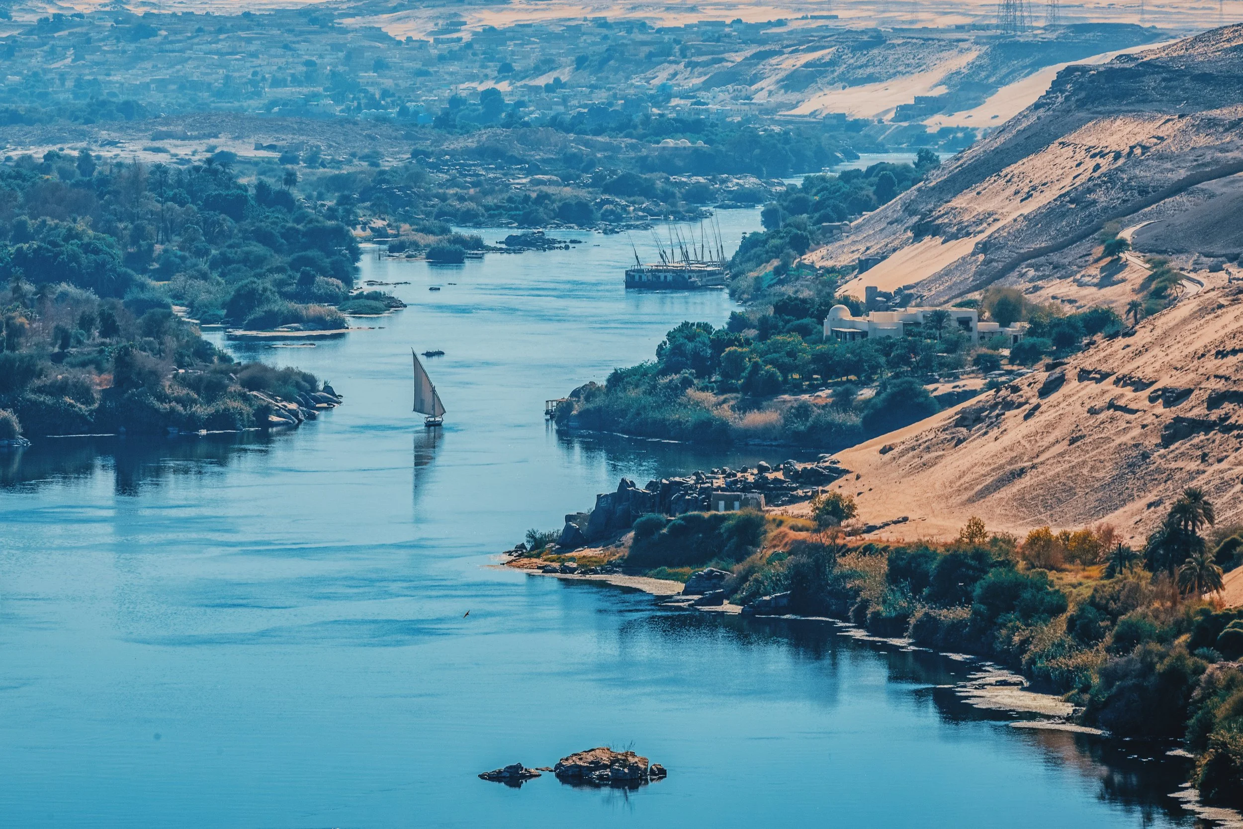 A felucca sailing down the Nile at Aswan between green islands and golden desert hills.