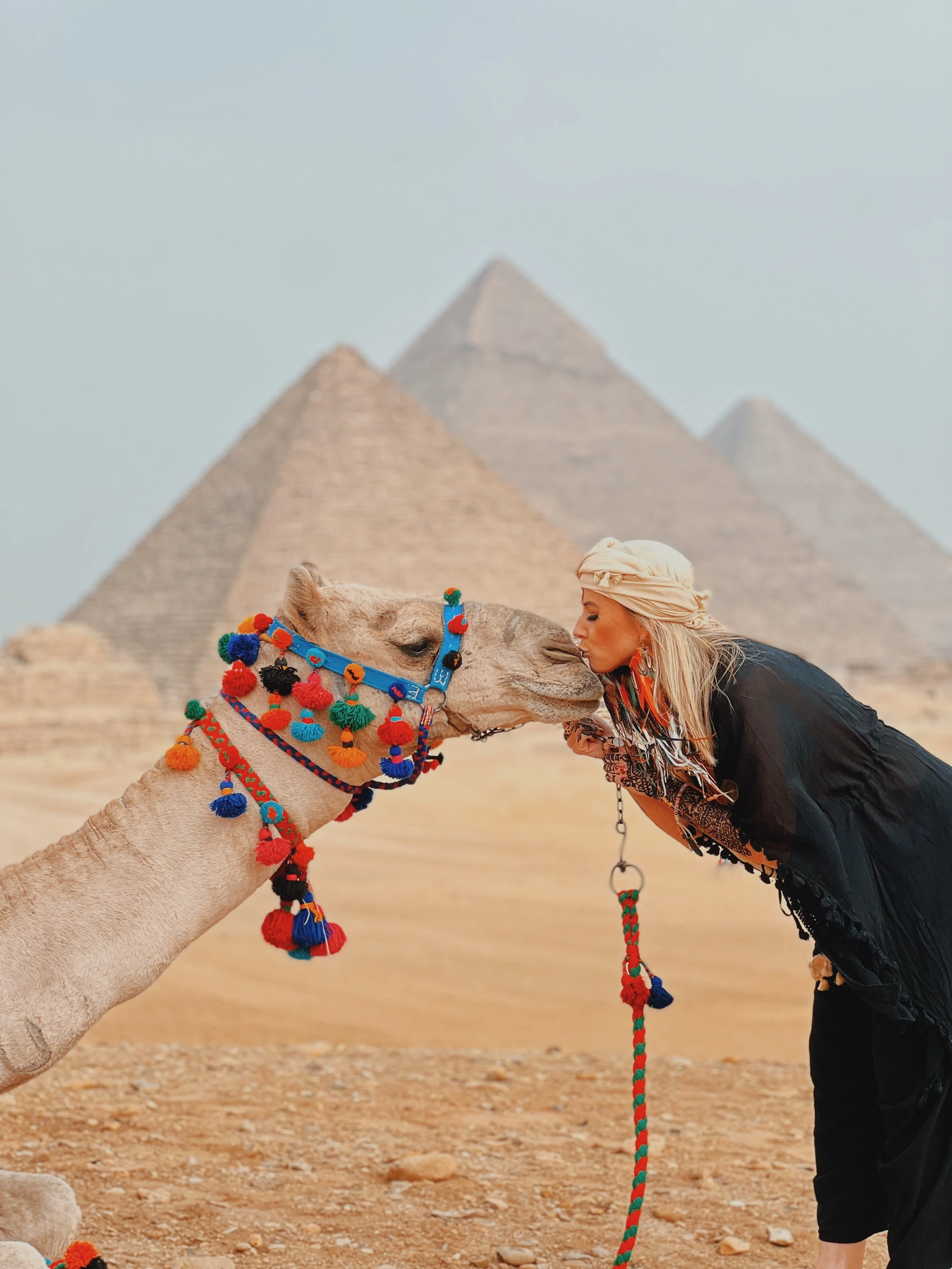 Jennifer in a cream headscarf leaning in to kiss a camel in a colorful tasseled halter, with the Pyramids of Giza behind them