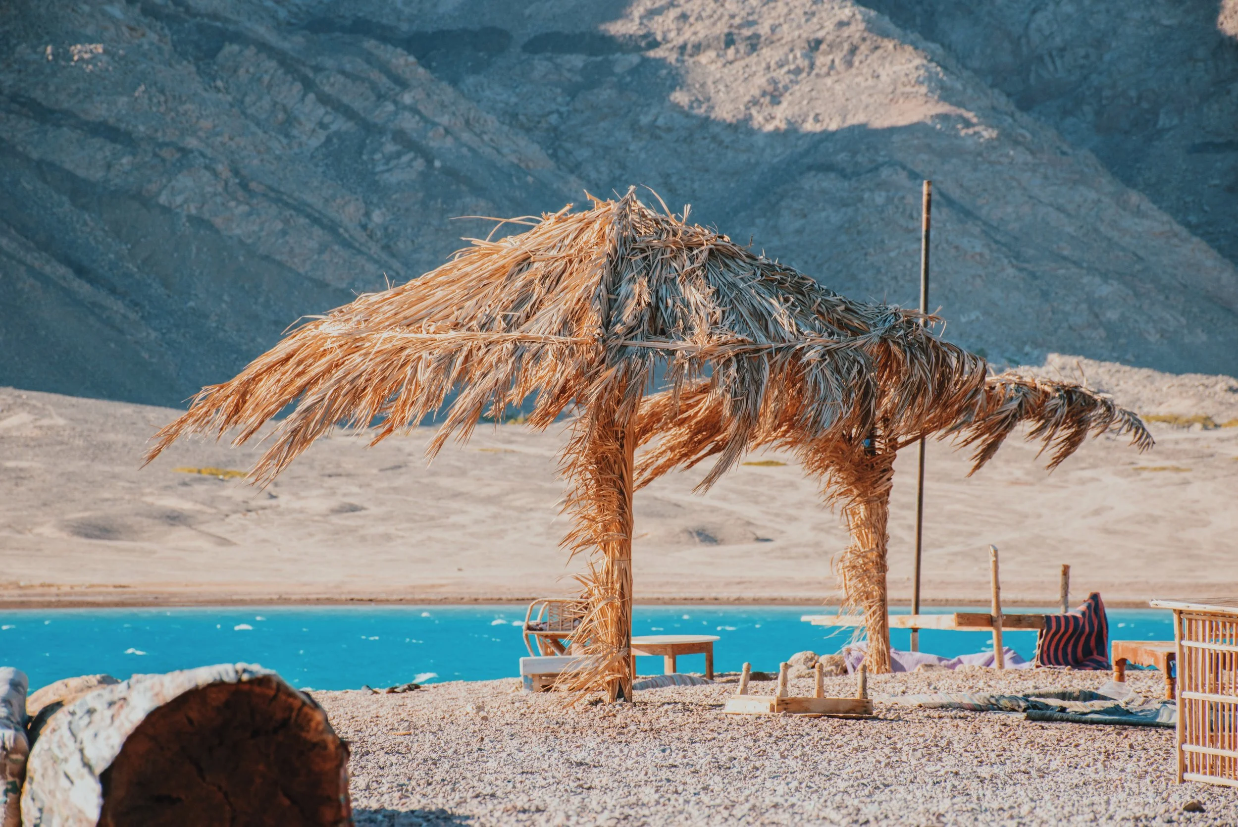 Palm-frond shades on the pebble beach of the Blue Lagoon near Dahab, with turquoise water and the Sinai mountains behind.
