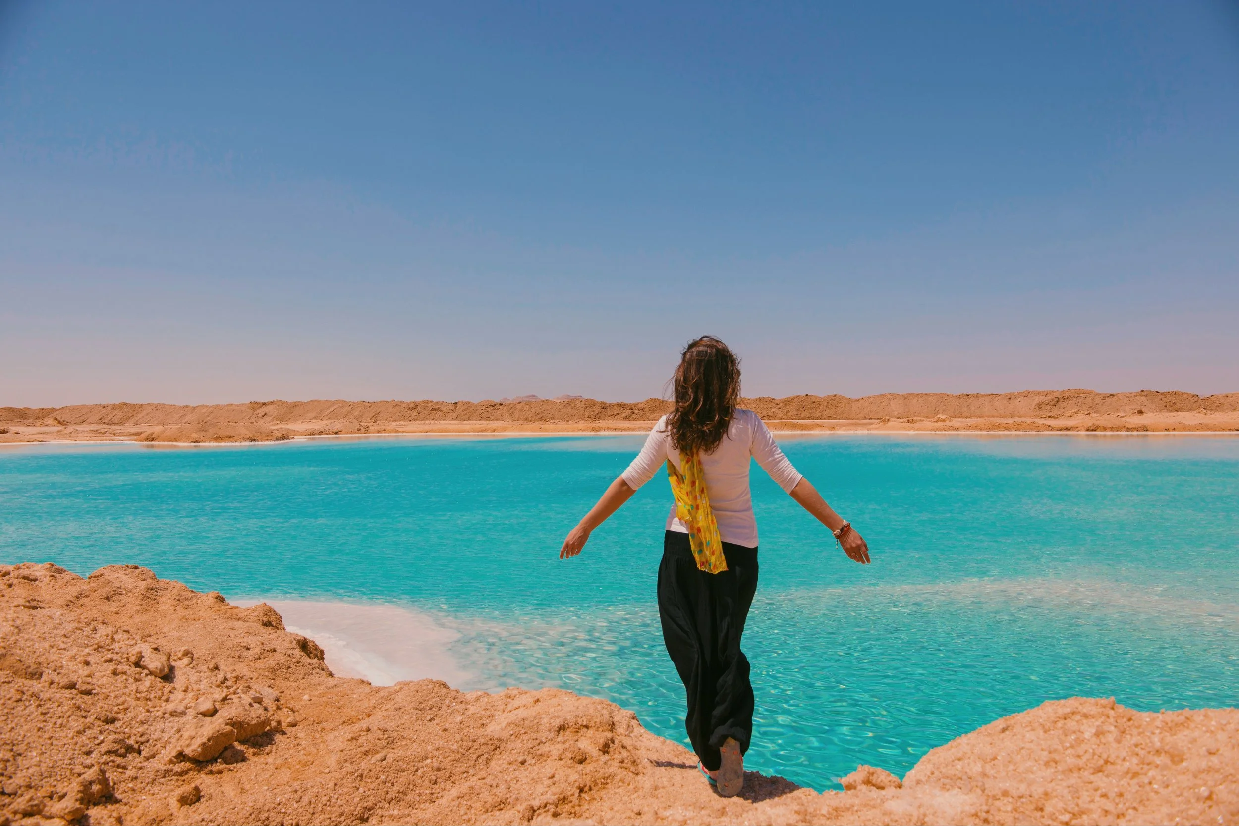 A woman with her arms open wide, standing at the edge of a turquoise salt lake in Siwa Oasis.