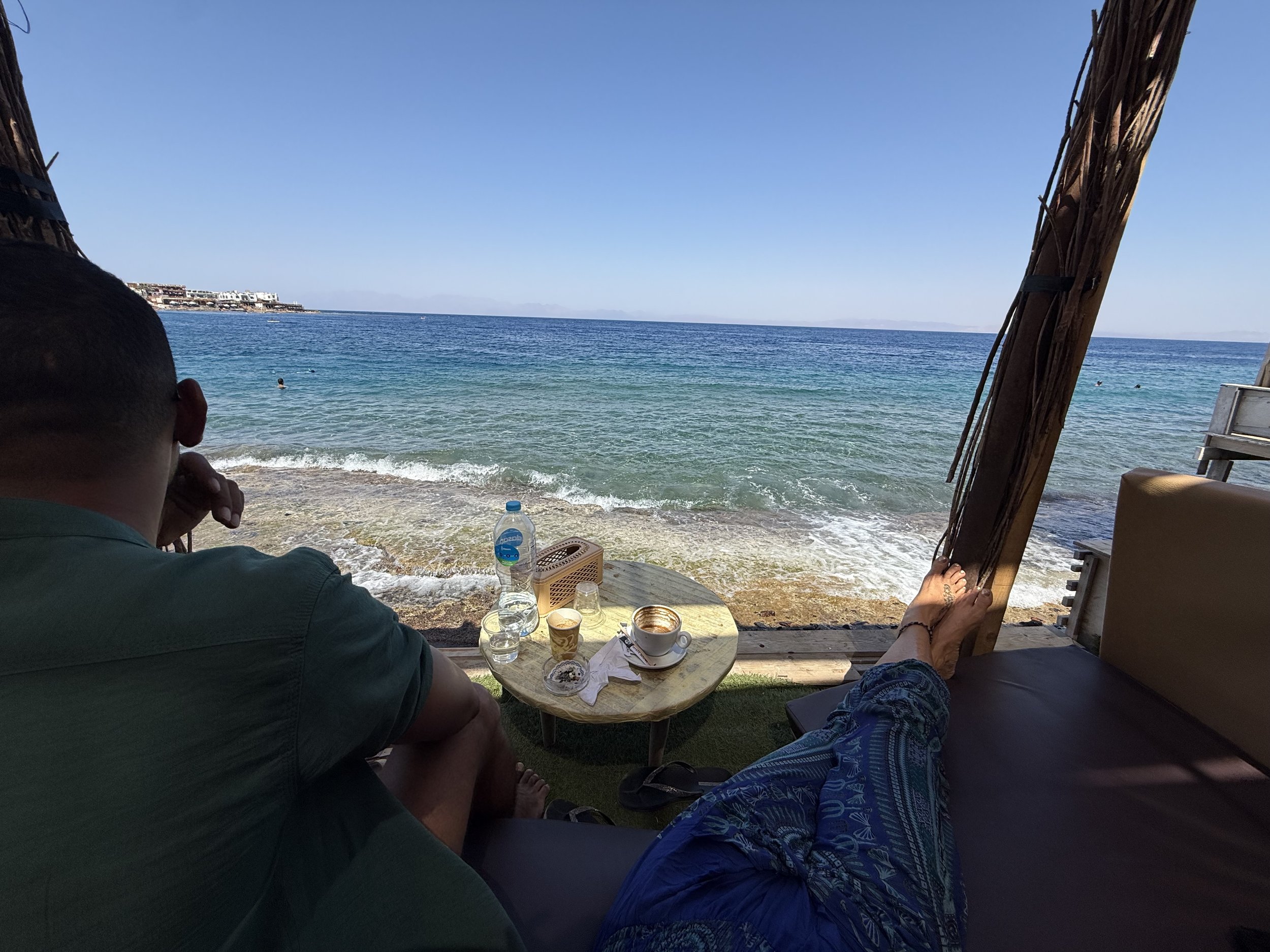 Bare feet up beside a Red Sea café table with coffee and water, looking out over the turquoise water of Dahab.