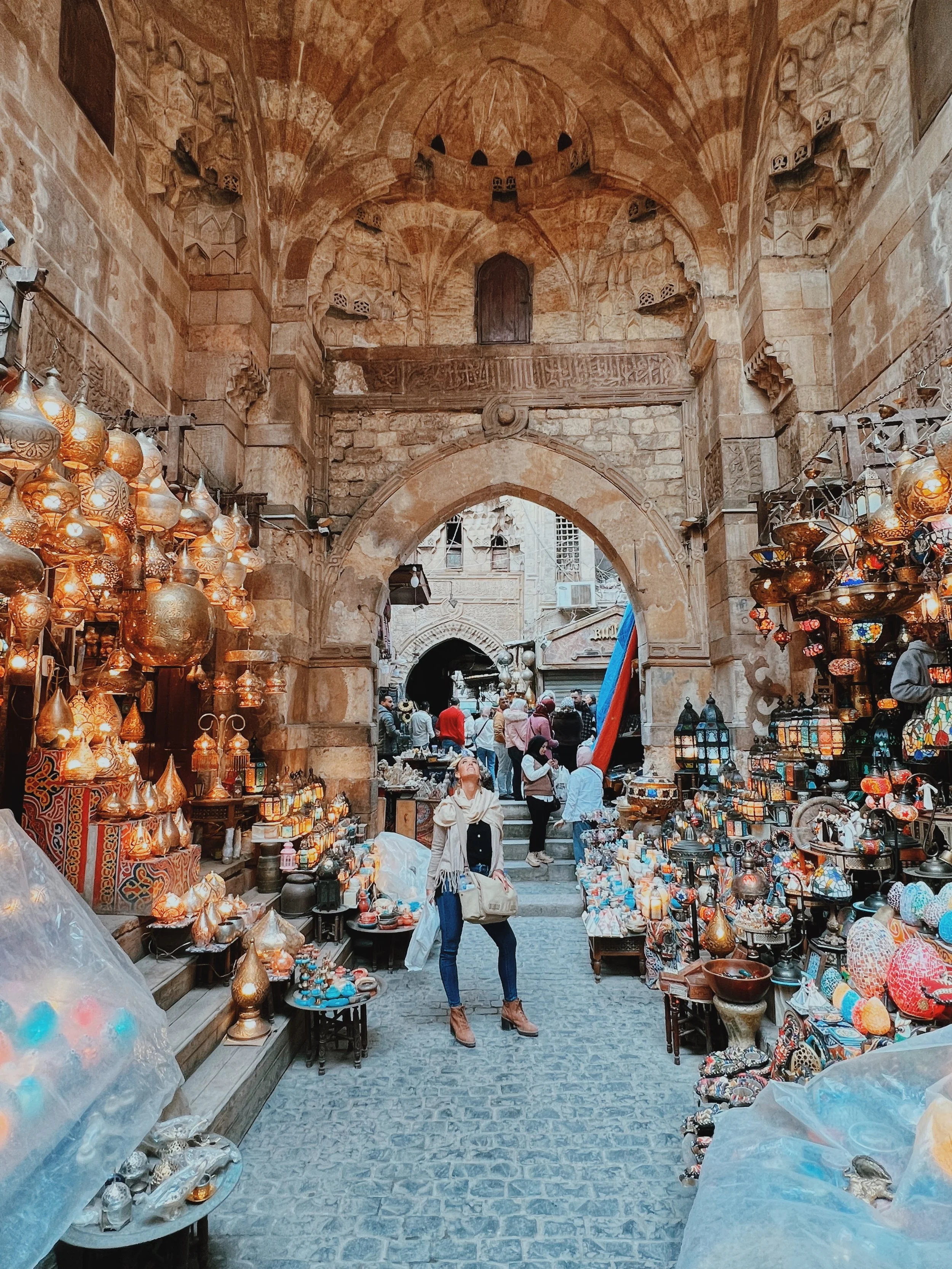 Jennifer gazing up at glowing brass lanterns beneath a medieval stone gateway in the Khan el-Khalili bazaar in Cairo