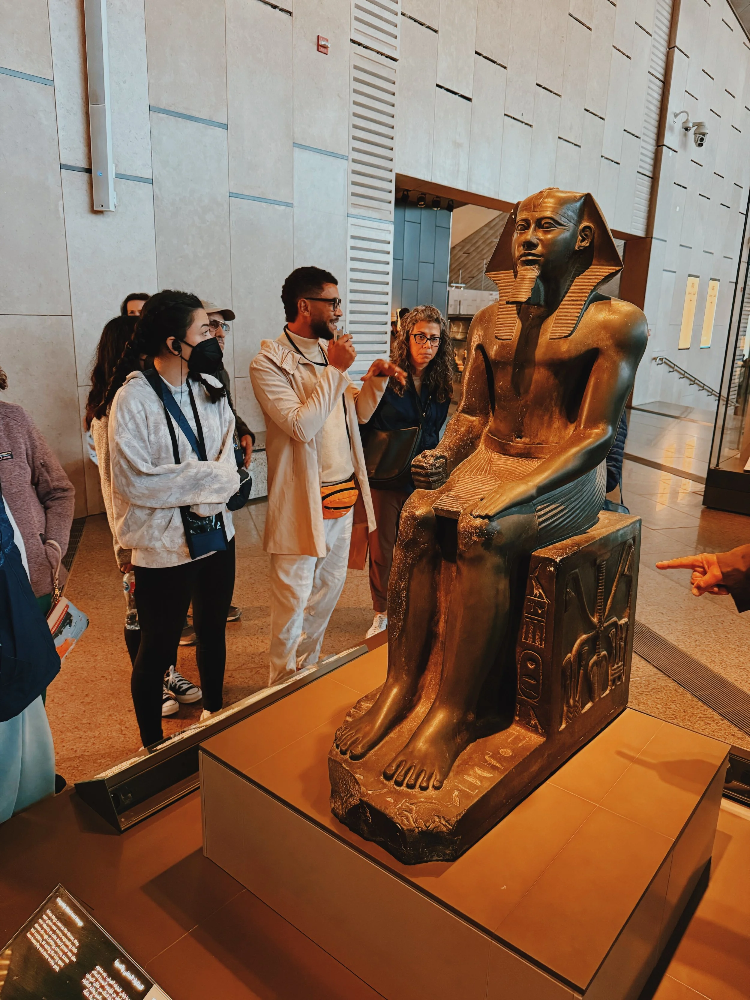Egyptologist Mostafa of Takemet Tours leading a group discussion beside a seated pharaoh statue at the Grand Egyptian Museum