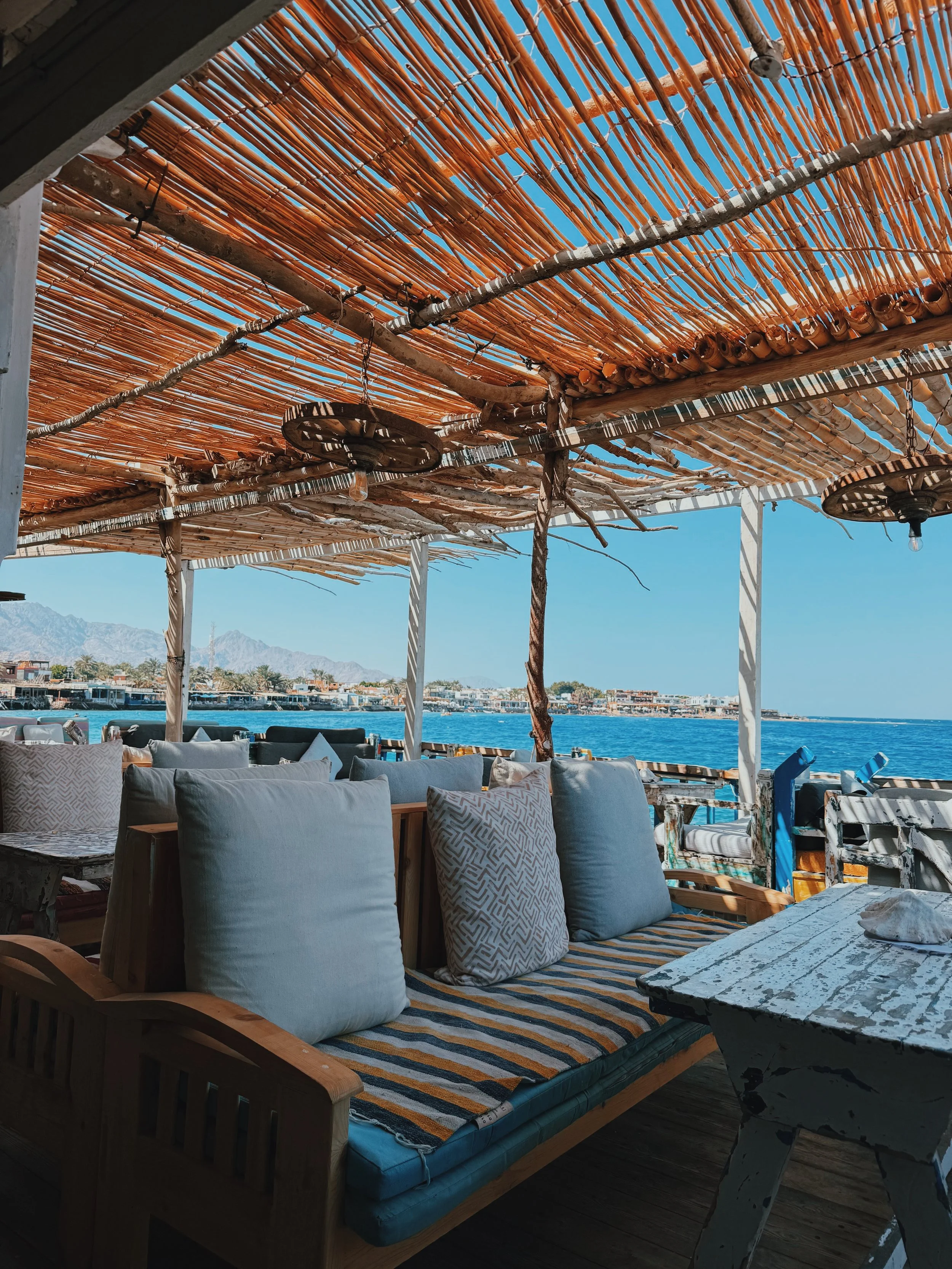 A reed-shaded café terrace with cushioned seating looking across the bay to Dahab and the Sinai mountains.