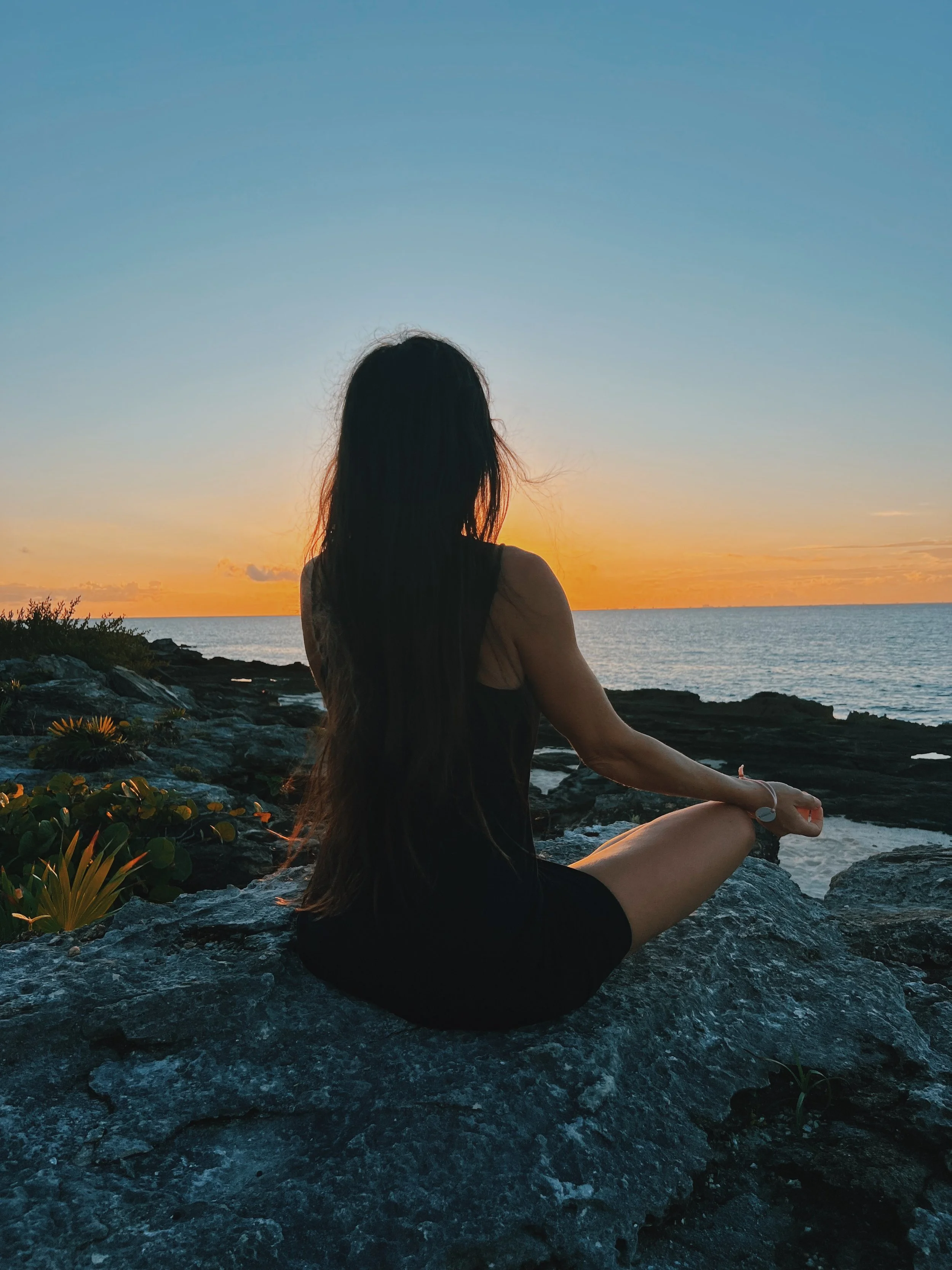 A woman meditating on the rocks by the sea at sunset