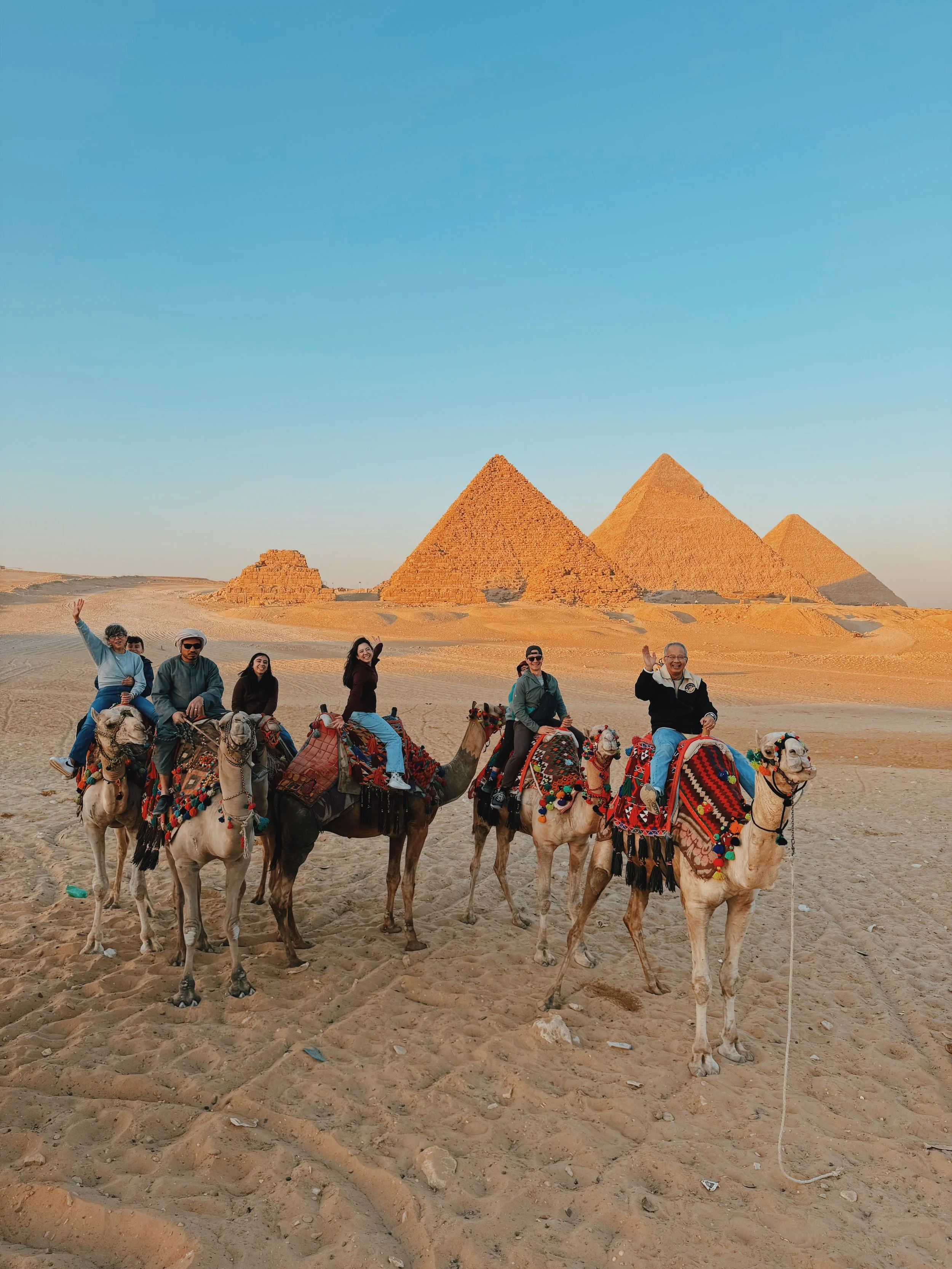 Takemet Tours guests waving from camels at sunset in front of the Pyramids of Giza