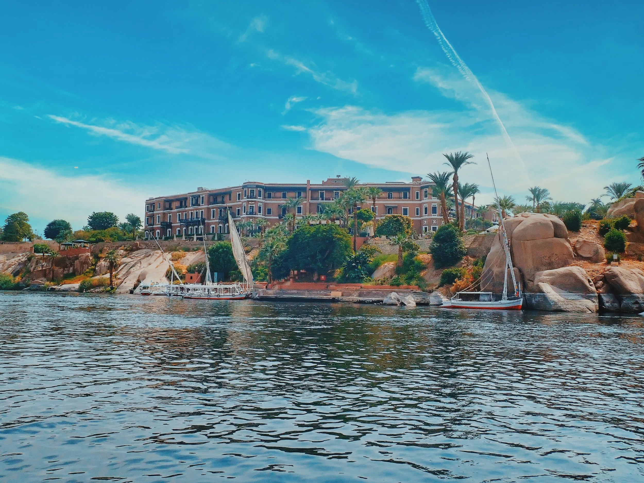 The historic Old Cataract Hotel overlooking the Nile and granite boulders in Aswan, with feluccas moored below.