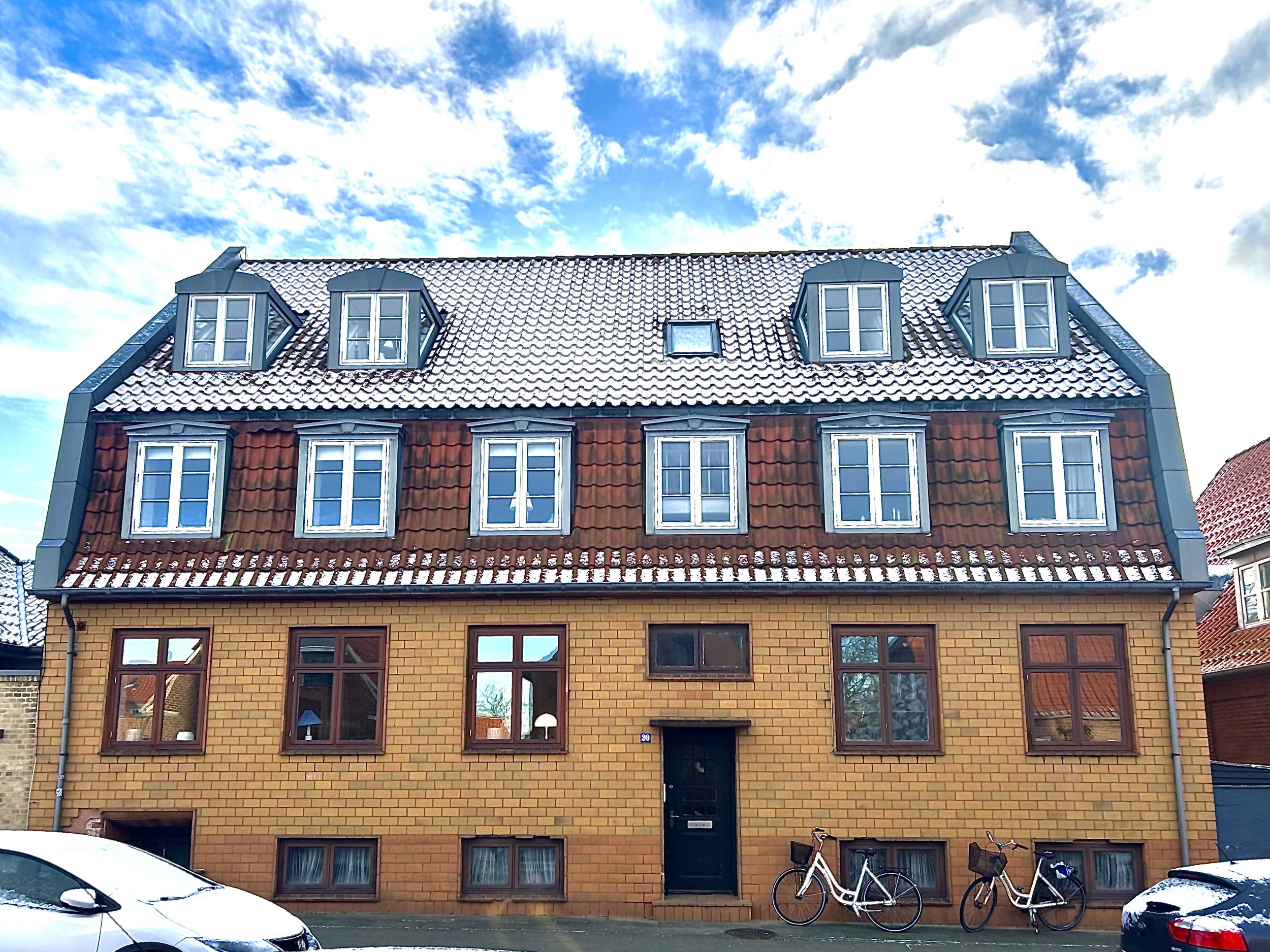 Et tre-ethus med gulbrune mursten, rødt tegltag og flere udvendige tagvinduer. Der er en sort hoveddør med trin udenfor, og der er affalds- og genbrugscontainere foran huset.