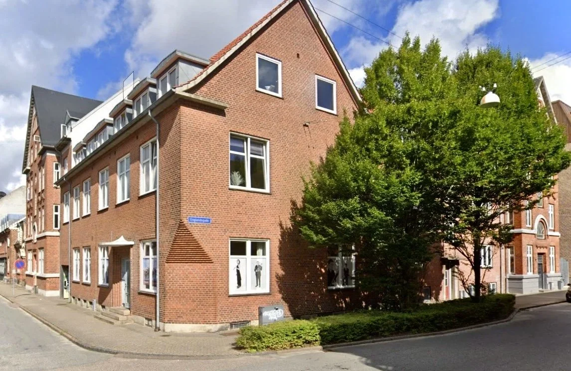 Et rødt murstensbyggeri på et hjørne med grønne træer foran. Gade- og husnumre synlige, en gade skilt med navnet Englandsvej.