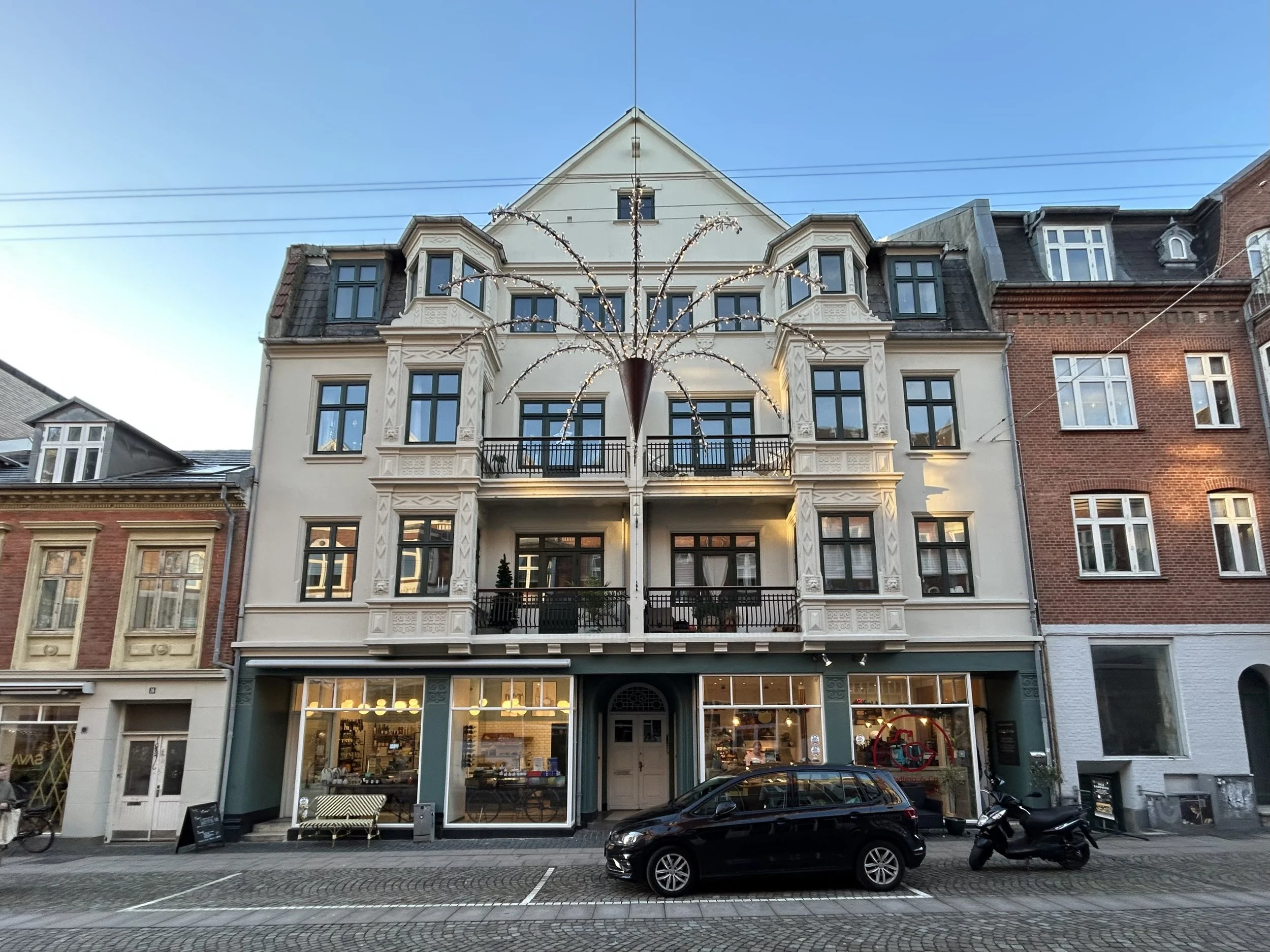 Ejendom i flere etager med klassisk arkitektur, store vinduer, balkon med sort gelænder og butik på stueplan.